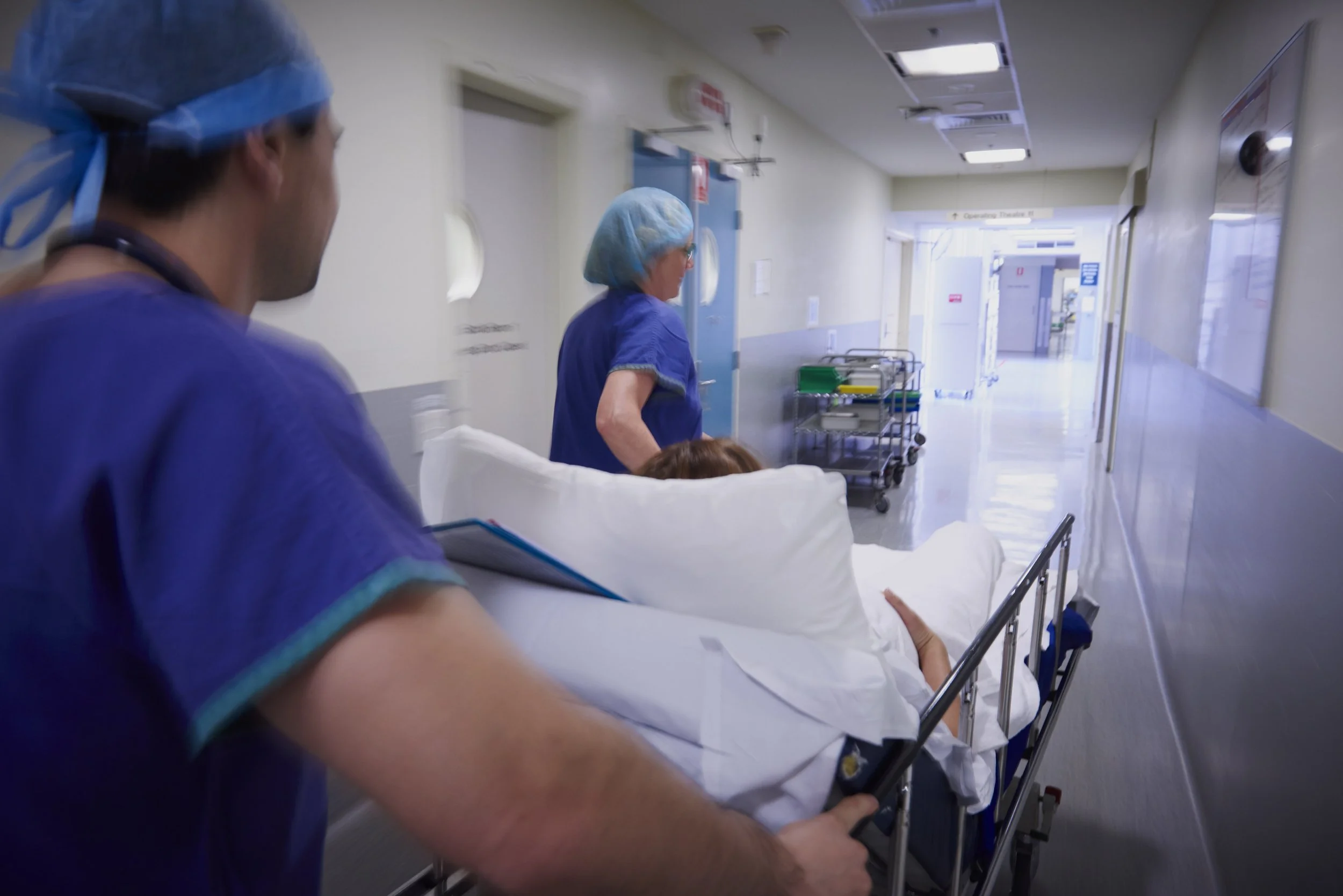 Medical staff members, wearing scrubs and surgical caps, move a patient on a hospital bed down a hallway in a hospital setting.