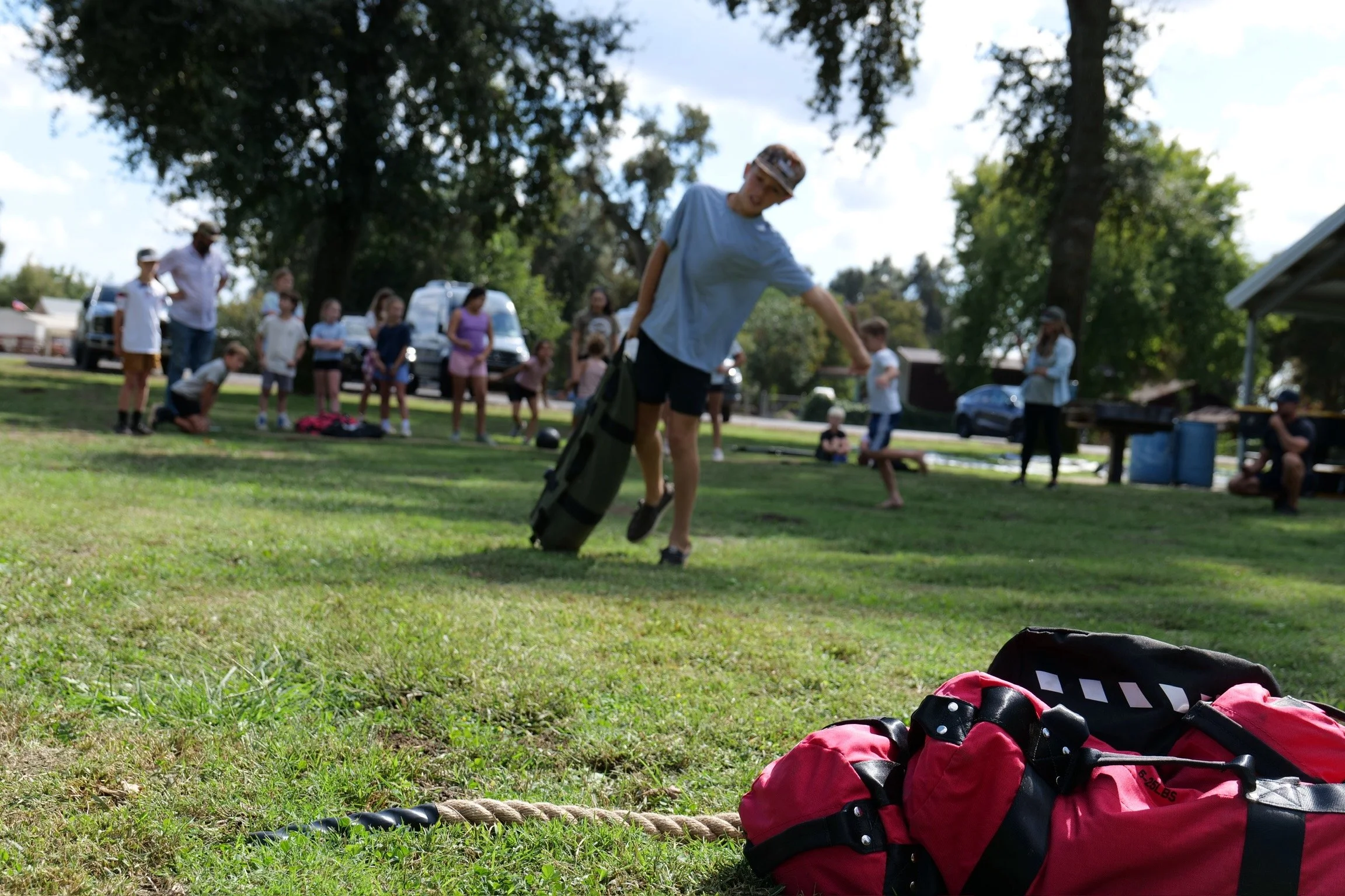 Group of children and adults outdoors, with a focus on a person in a blue shirt and cap near the center, as they prepare for activity, with gym bags and ropes on the grass in the foreground, and cars and trees in the background.