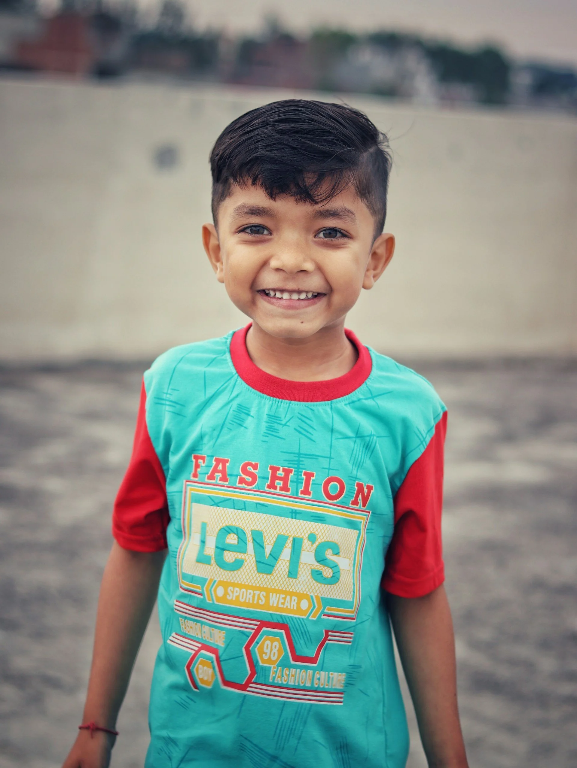 A young boy with dark hair smiling outdoors, wearing a turquoise and red Levi's shirt.