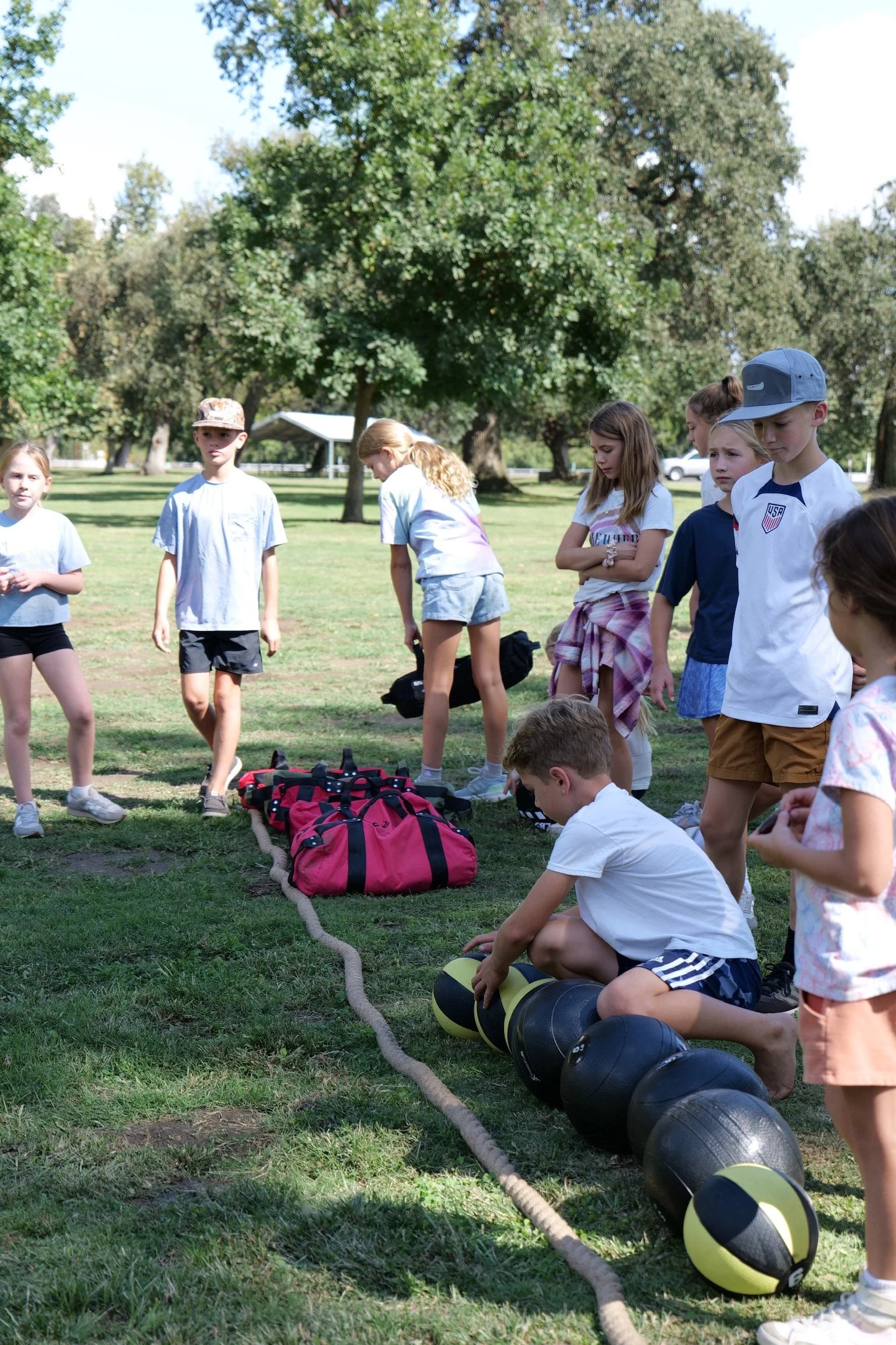A group of children outdoors on a grassy field, preparing for a fitness activity, with punching bags, gym bags, and a rope on the ground, surrounded by trees.