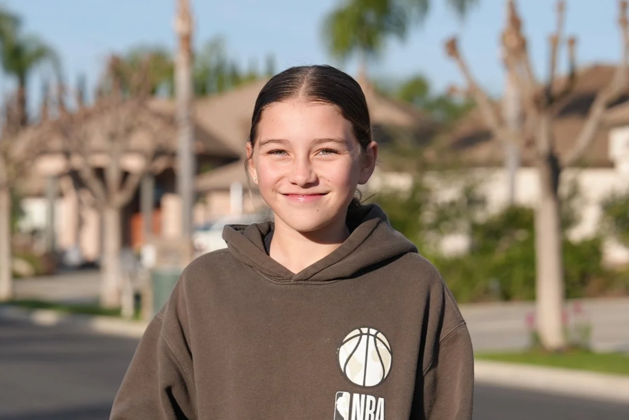 A young girl with curly hair, wearing a headband, earrings, and a patterned dress, standing outdoors with buildings blurred in the background.