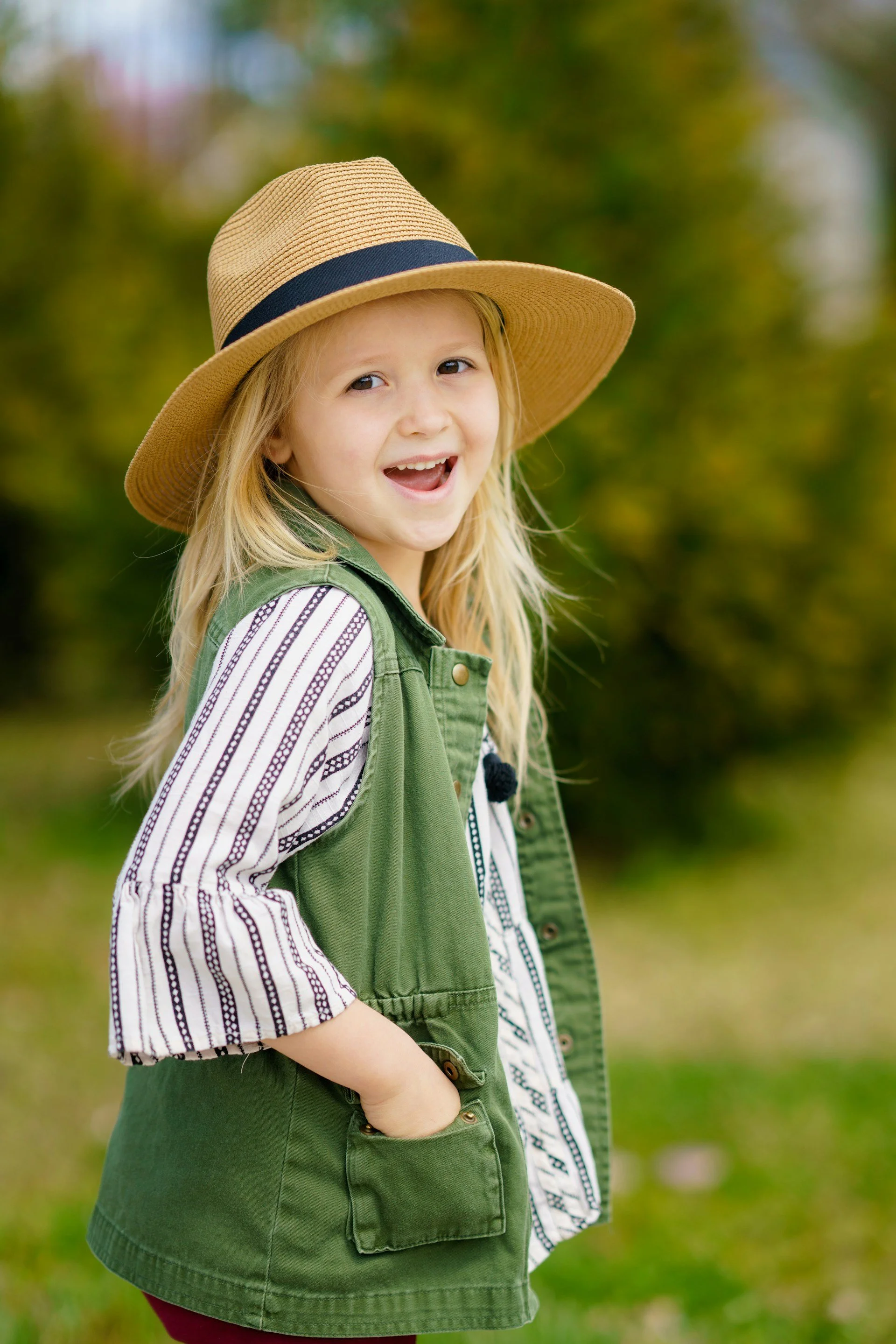 A young girl with long blonde hair wearing a beige wide-brimmed hat, a green vest, and a striped white and black shirt outdoors with blurred trees in the background.