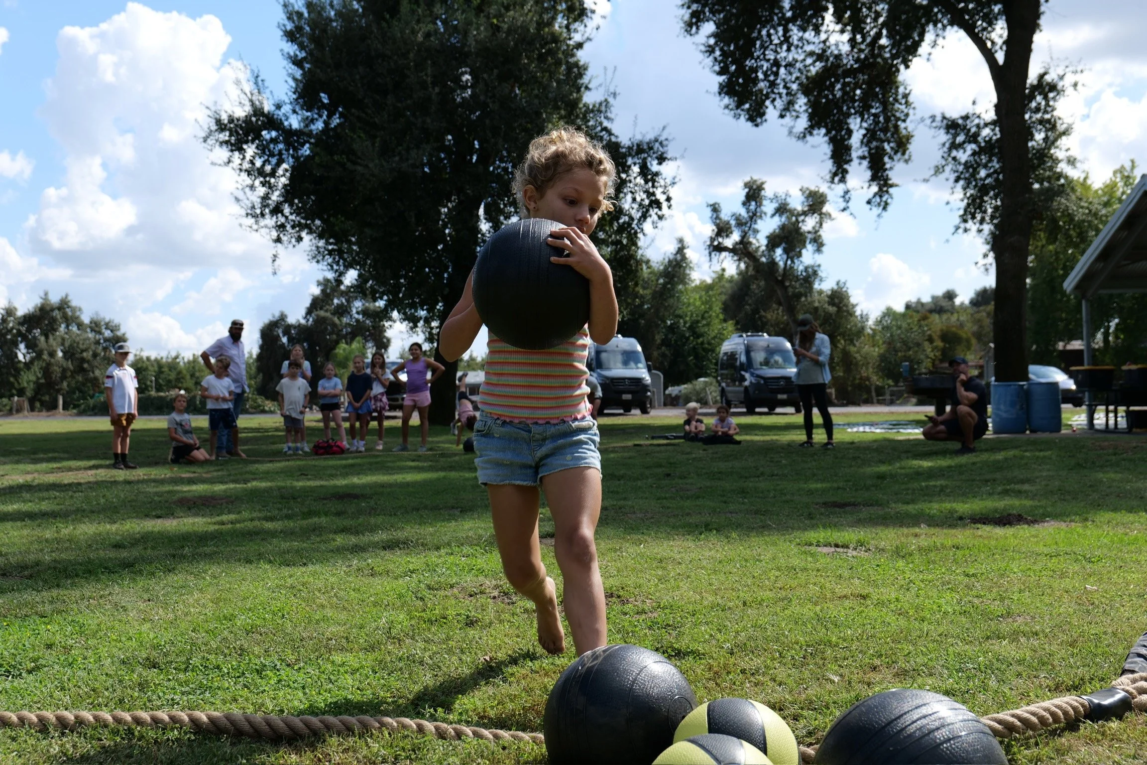 Young girl lifting a black medicine ball in a park during an outdoor event, with a group of children and adults in the background under a partially cloudy sky.