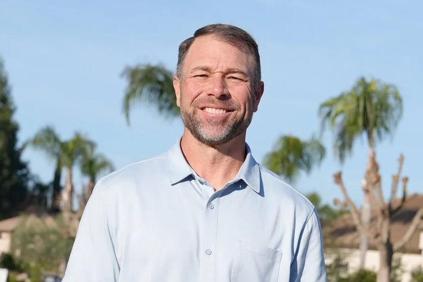 A young man with glasses and a beard standing outdoors during sunset, smiling slightly at the camera.