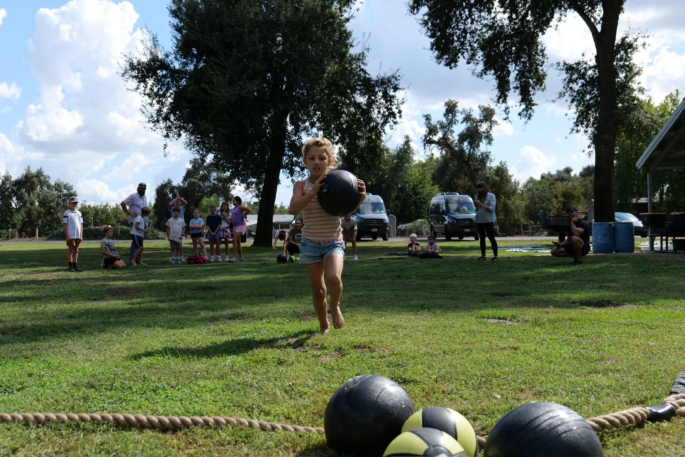 A young girl runs towards a black medicine ball on a grassy field during an outdoor group activity or sports event.