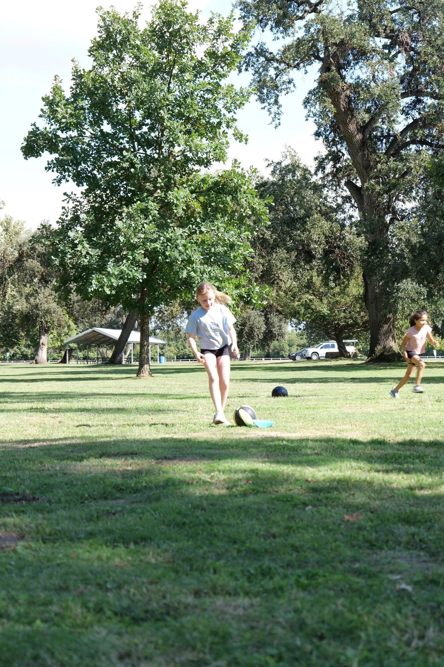 Children playing football in a park on a sunny day, surrounded by large trees and open grassy area.
