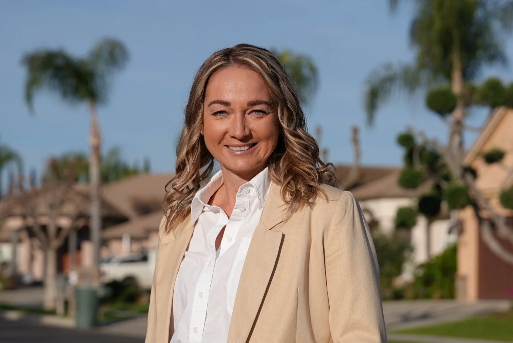 Smiling woman with dark shoulder-length hair, wearing a black fuzzy sweater and light-colored pants, sitting on a striped blanket on a beige couch with beige pillows and a plain cream wall behind her.