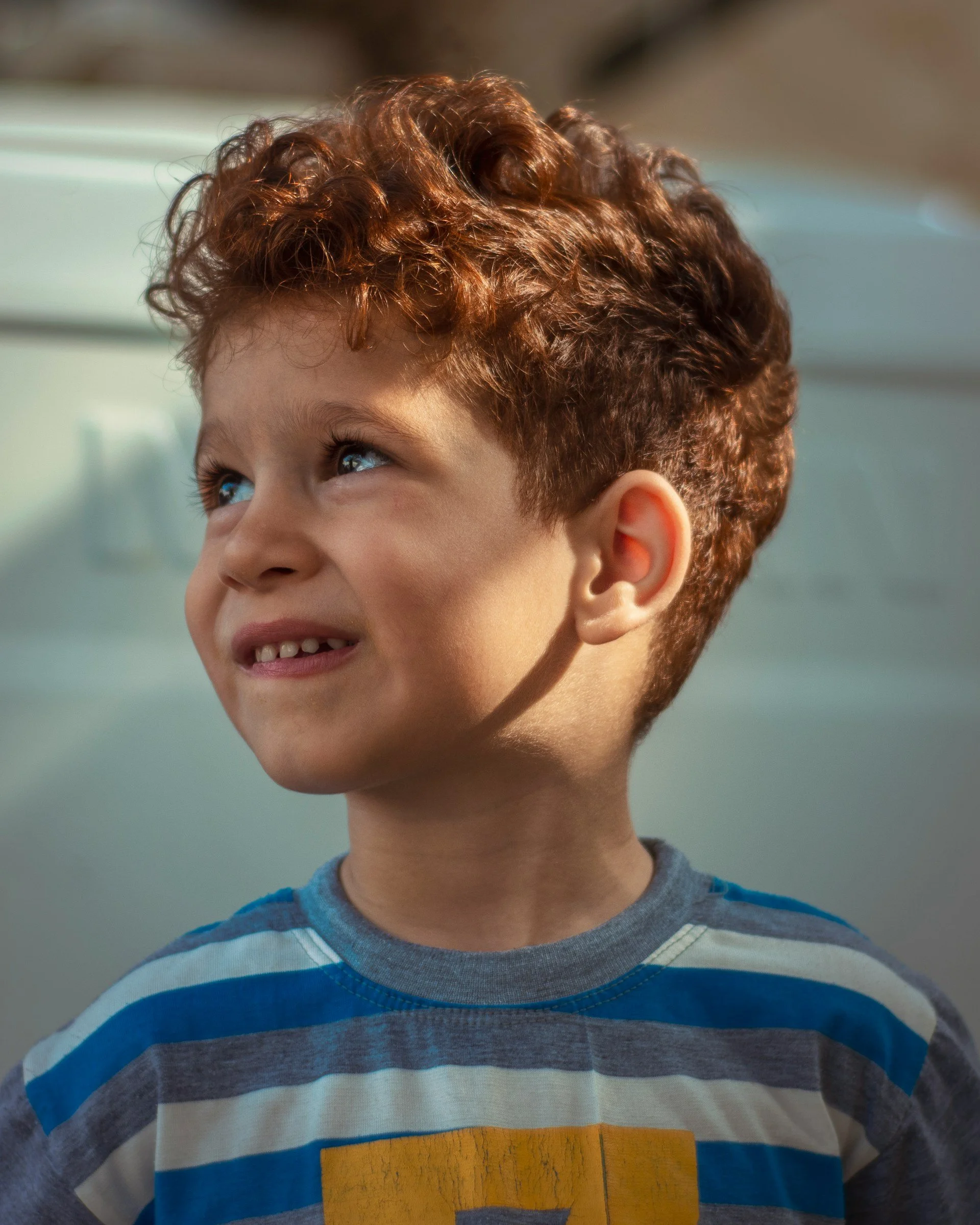 A young boy with curly red hair and blue eyes looking upward, smiling slightly, wearing a striped blue and gray shirt.