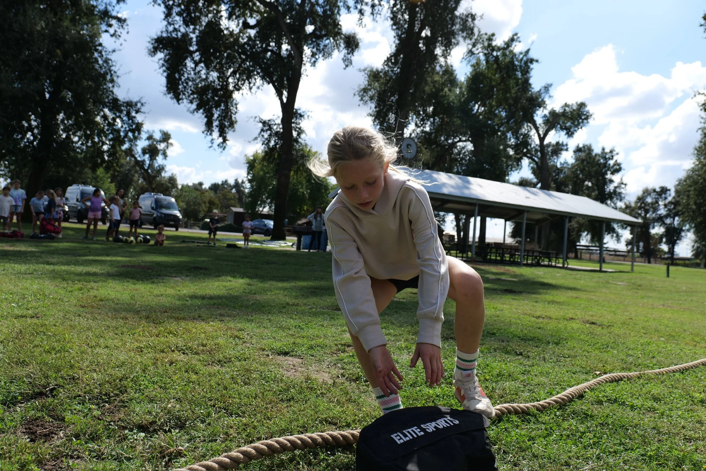 Young girl tying her shoelace on a grassy field during a sports event at a park with trees, a pavilion, and people in the background.