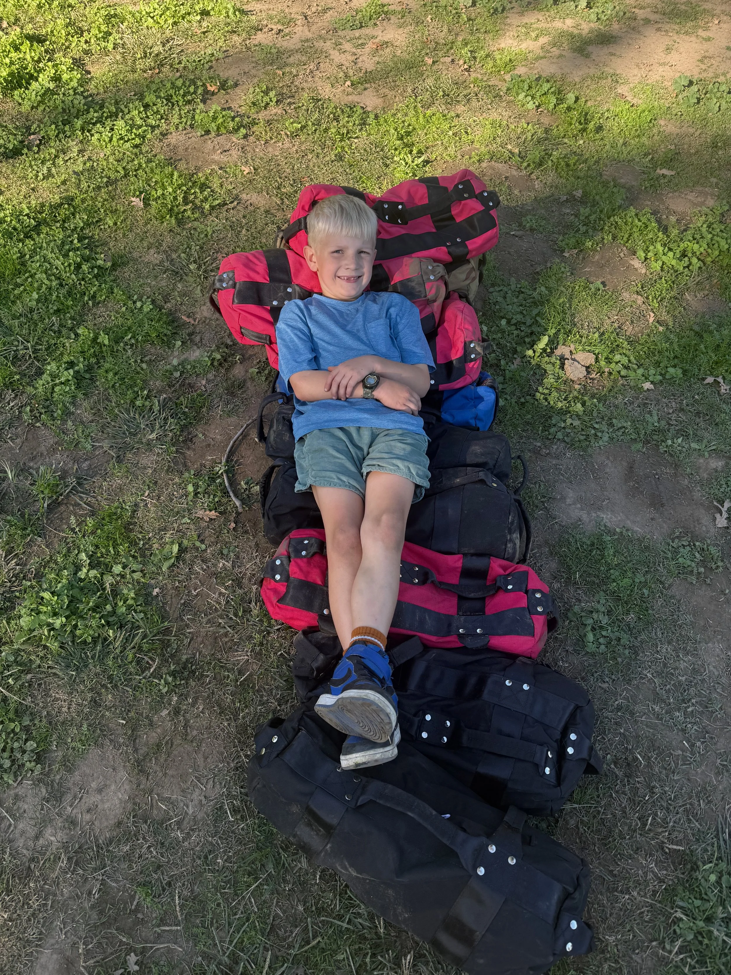 A smiling young boy with blond hair and a blue shirt, lying on top of multiple stacked backpacks on the ground outdoors, surrounded by grass and dirt.