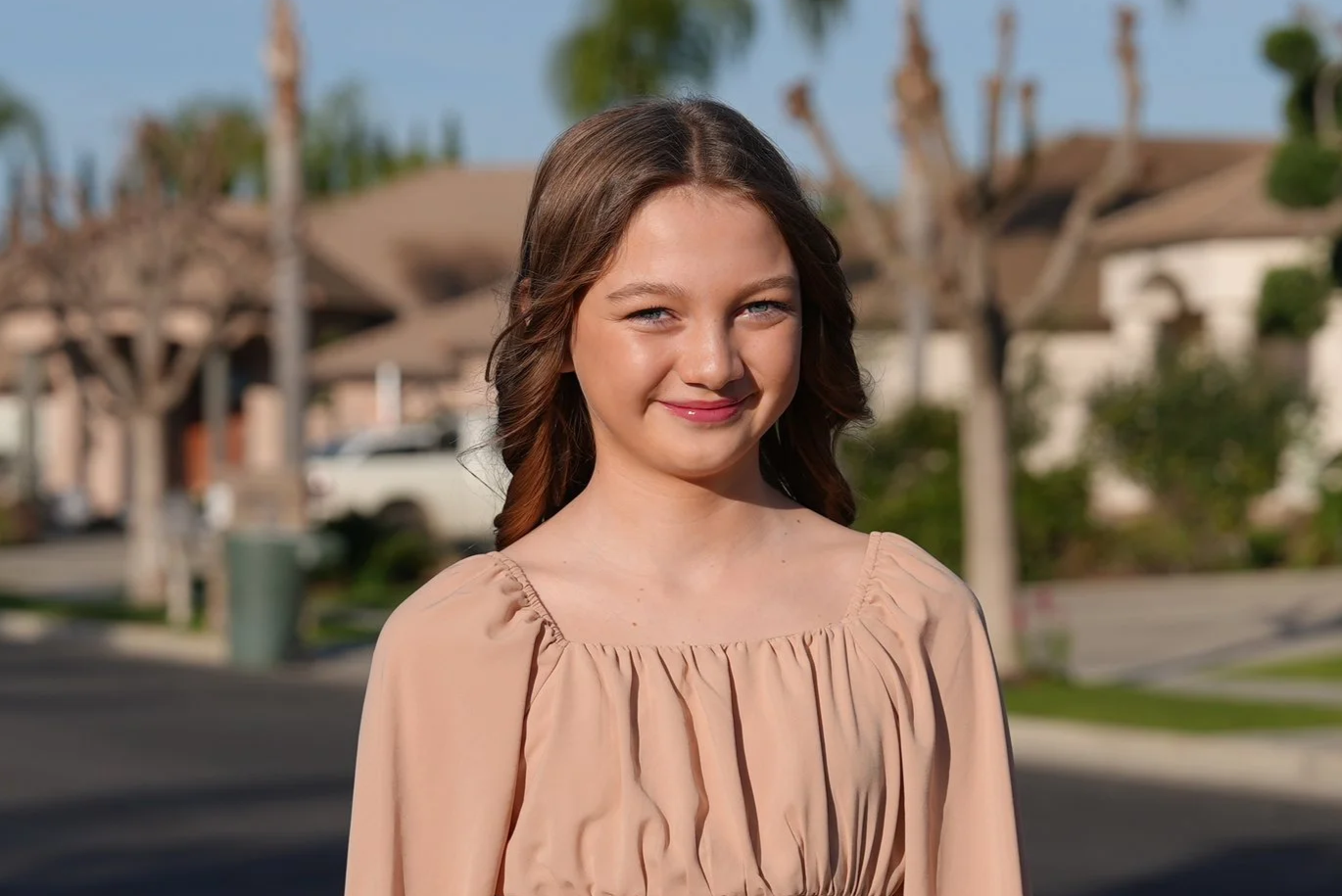 A young girl with long blonde hair wearing a beige wide-brimmed hat, a green vest, and a striped white and black shirt outdoors with blurred trees in the background.