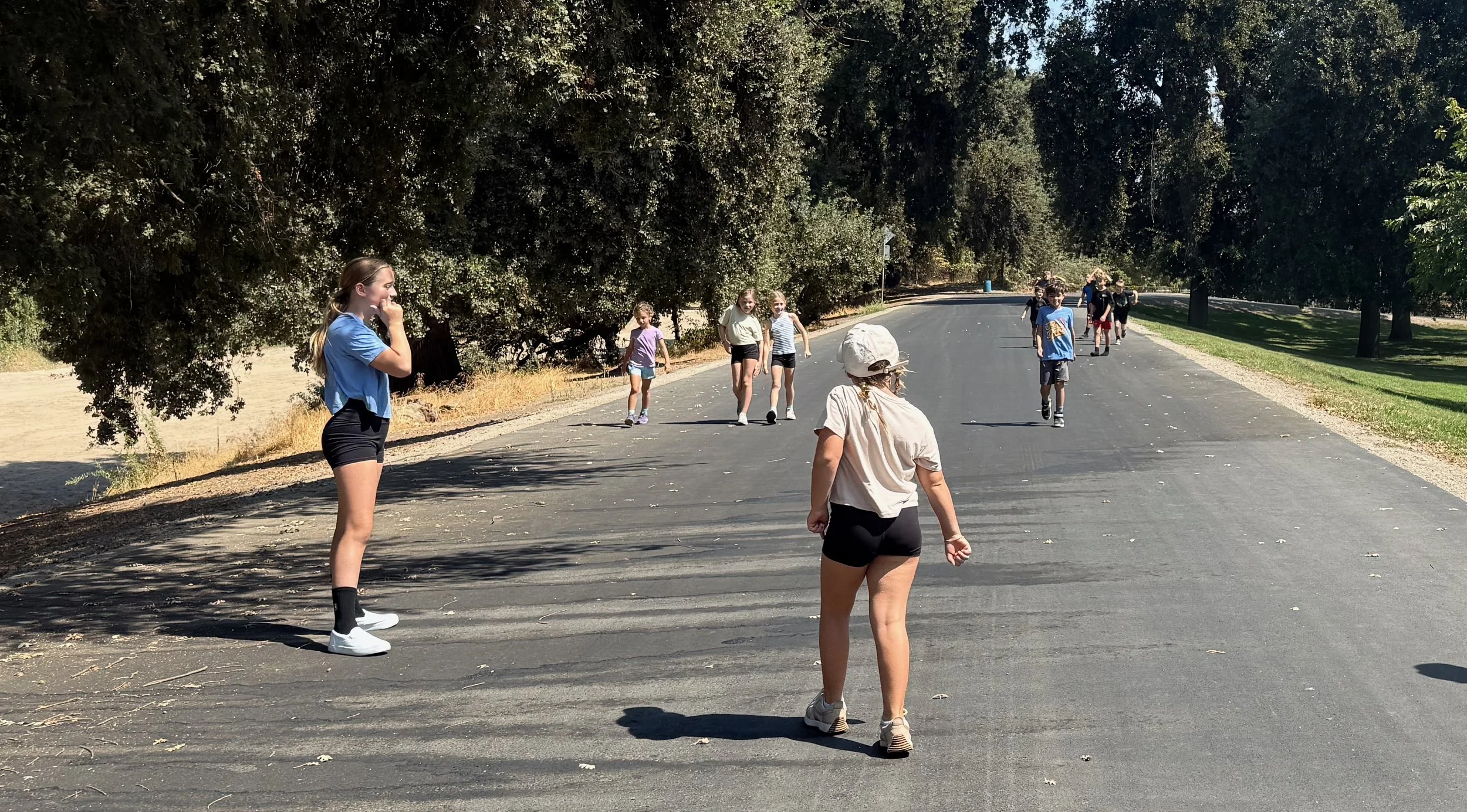 A group of children walking on a paved path in a park, with trees on both sides. One girl in the foreground is wearing a beige shirt, black shorts, and a white hat, while another girl in a blue shirt and black shorts stands to the left. In the background, several children are walking and talking, under a sunny sky.