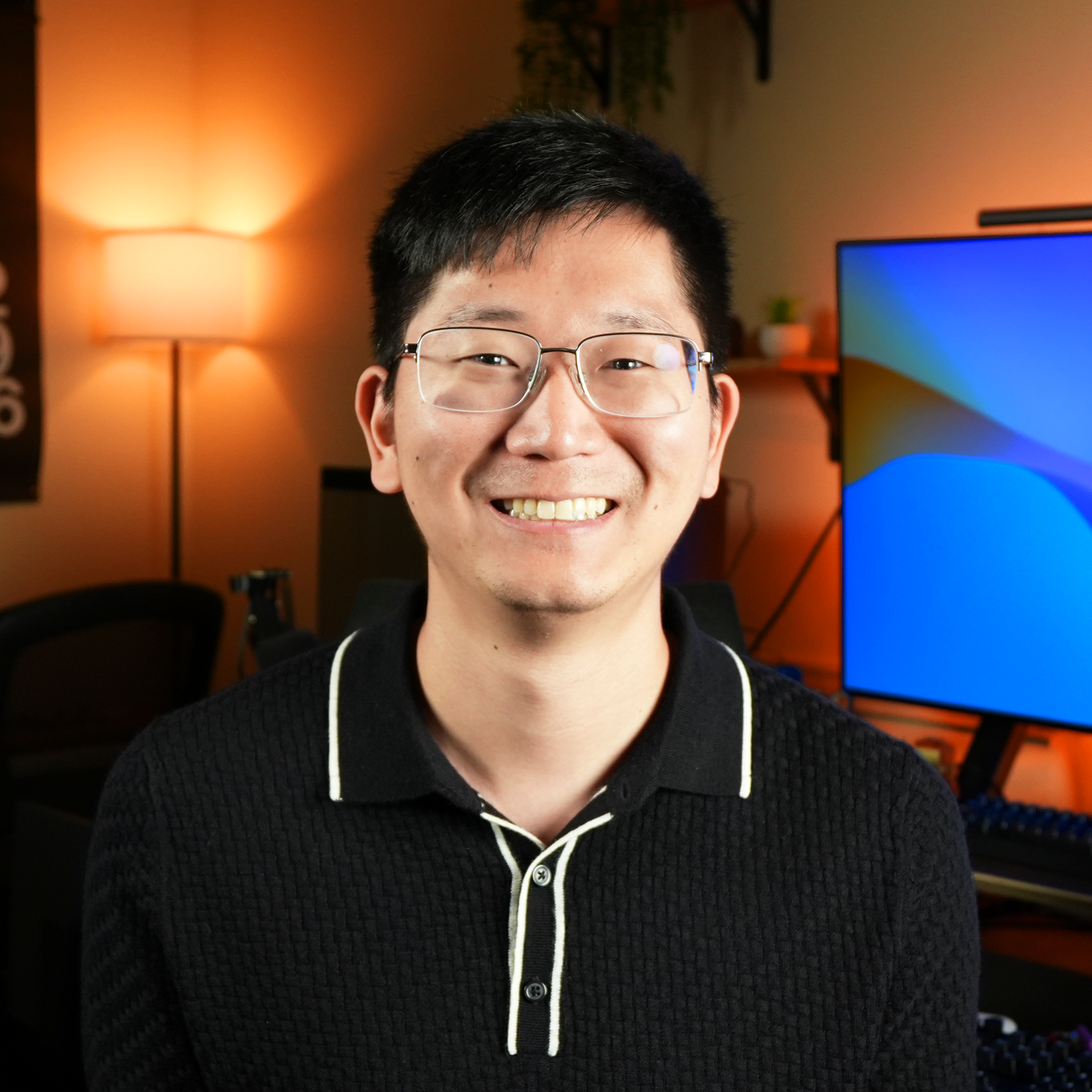 A smiling man with glasses in a room with warm lighting, a desk, a computer monitor, and a lamp in the background.