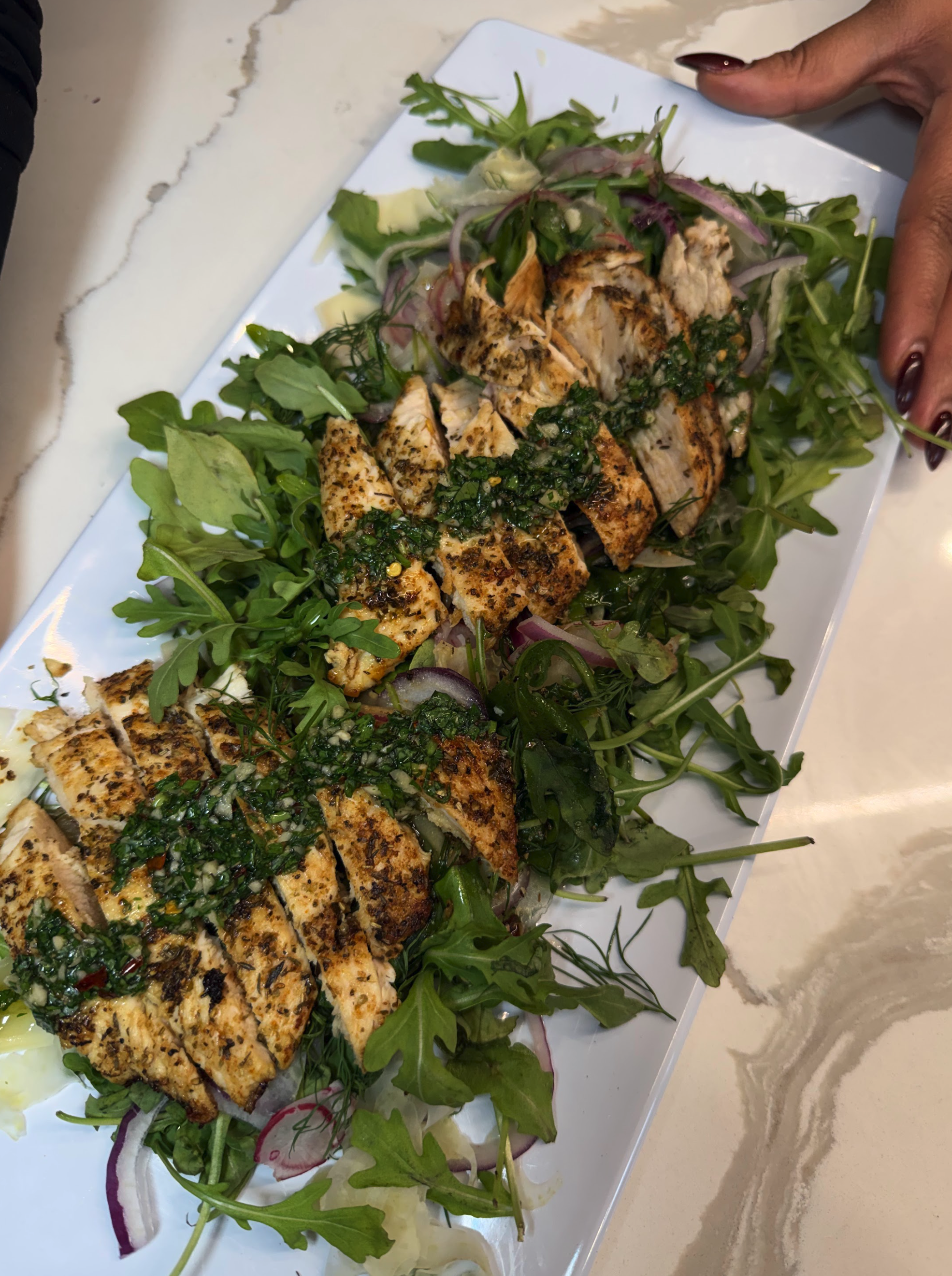 Grilled chicken sliced and topped with herbs, served on a bed of mixed greens and vegetables on a white rectangular plate