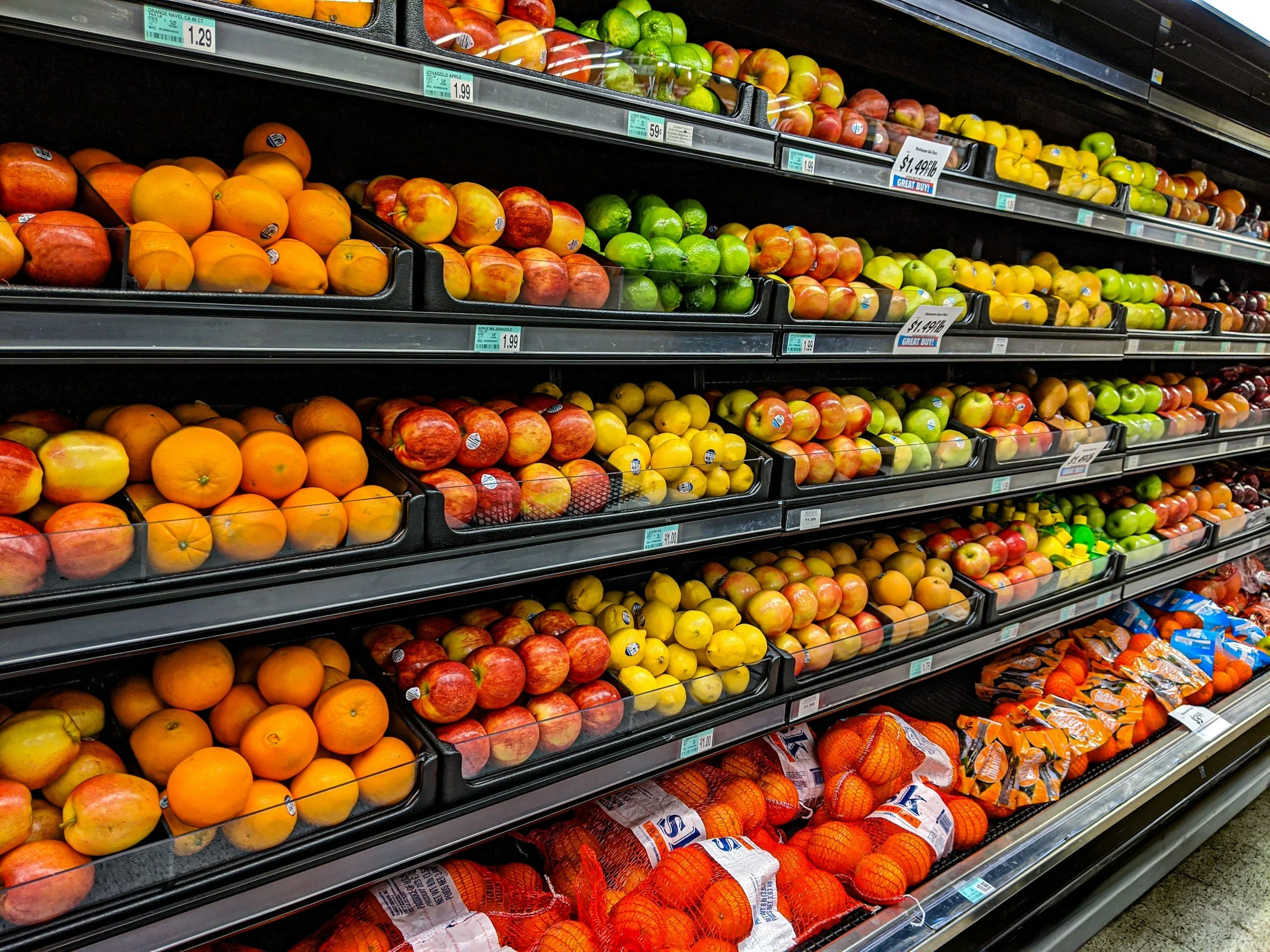 The image shows multiple shelves in a grocery store filled with apples, oranges, and other fruits packaged in plastic trays and mesh bags.
