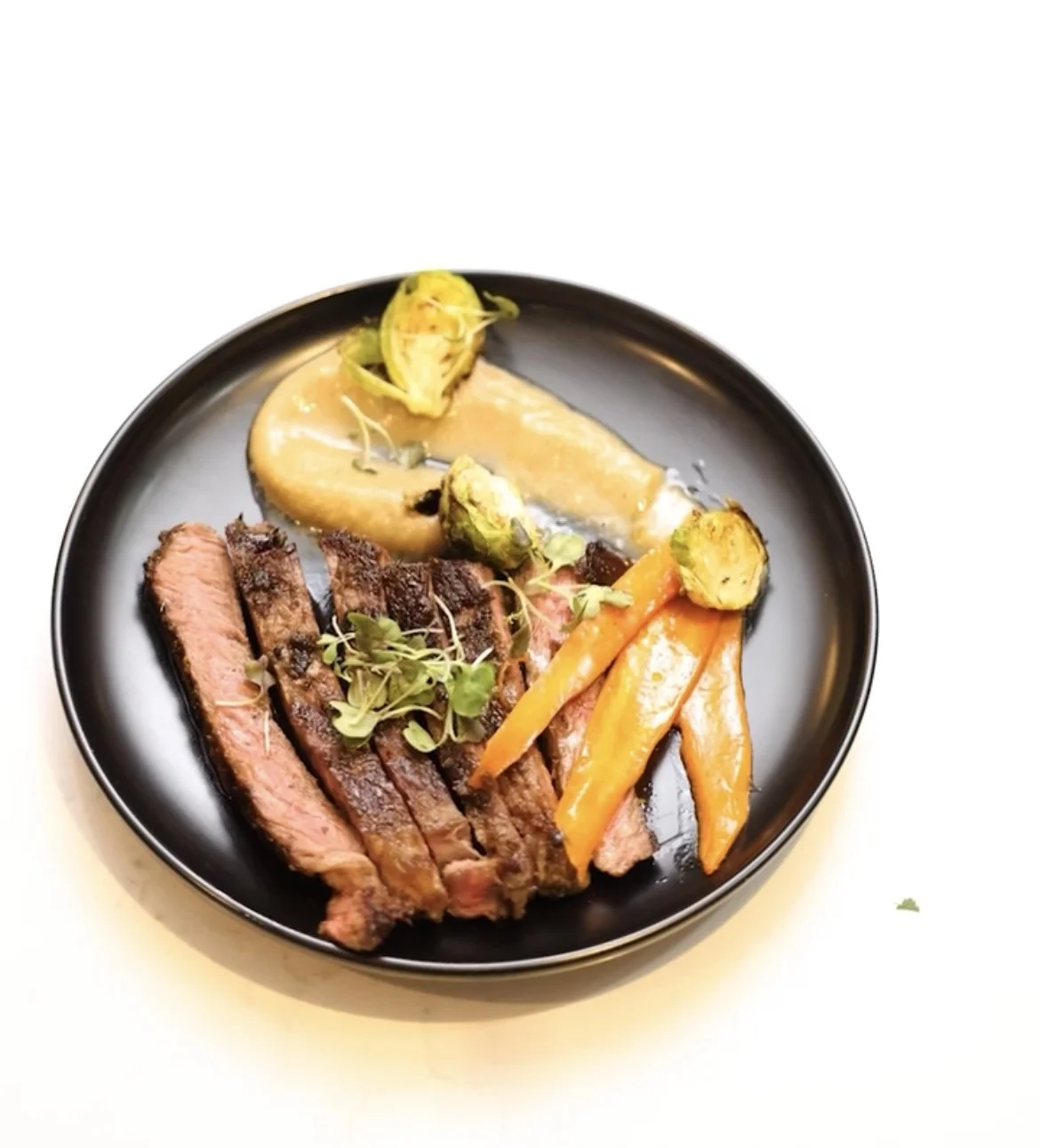 A black plate with sliced steak, roasted vegetables including carrots, Brussels sprouts, and a baked potato topped with cheese and sour cream, garnished with microgreens.