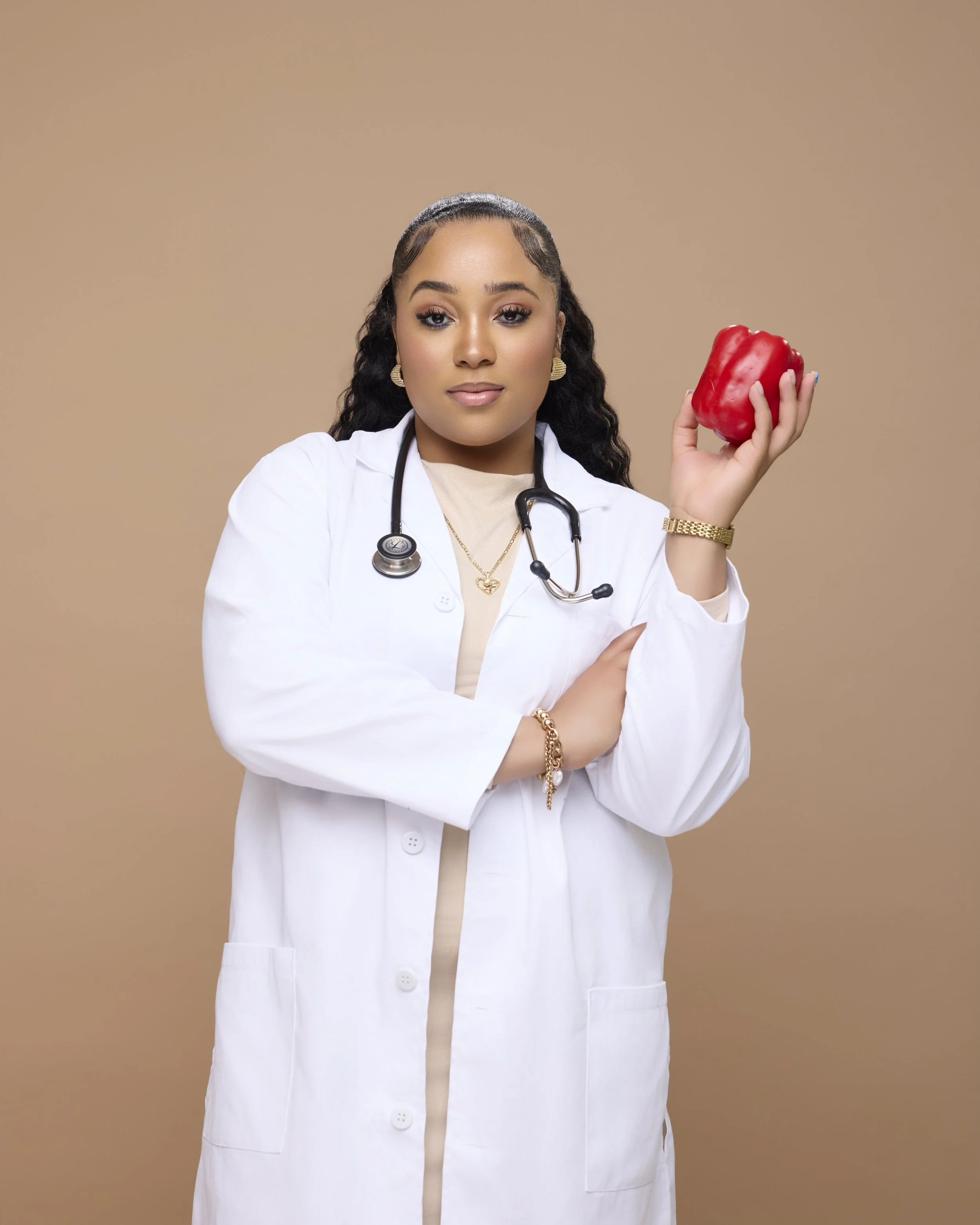 A female doctor wearing a white lab coat and stethoscope, holding a red bell pepper, against a beige background.
