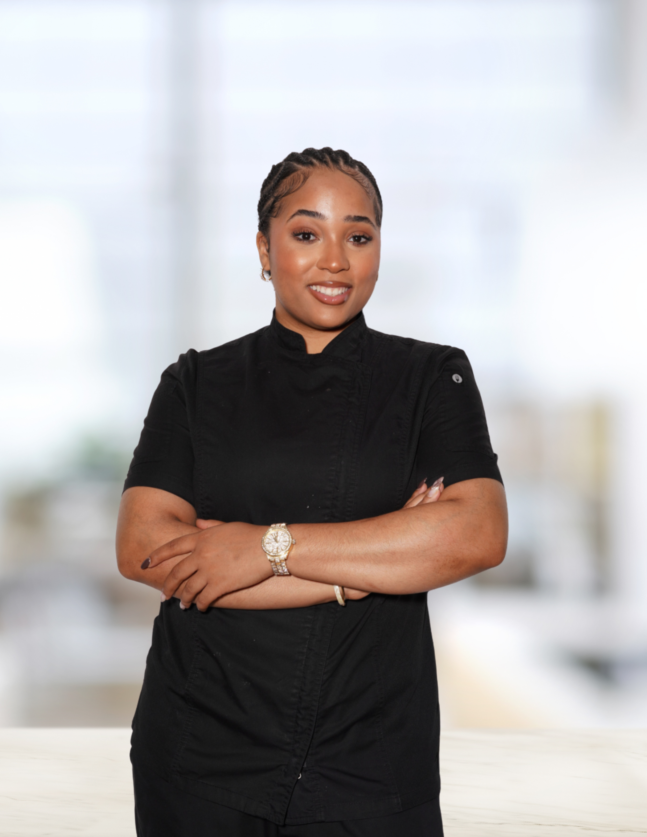 Portrait of a woman with braided hair, wearing a black top, crossing her arms, and smiling in front of a blurred indoor background.