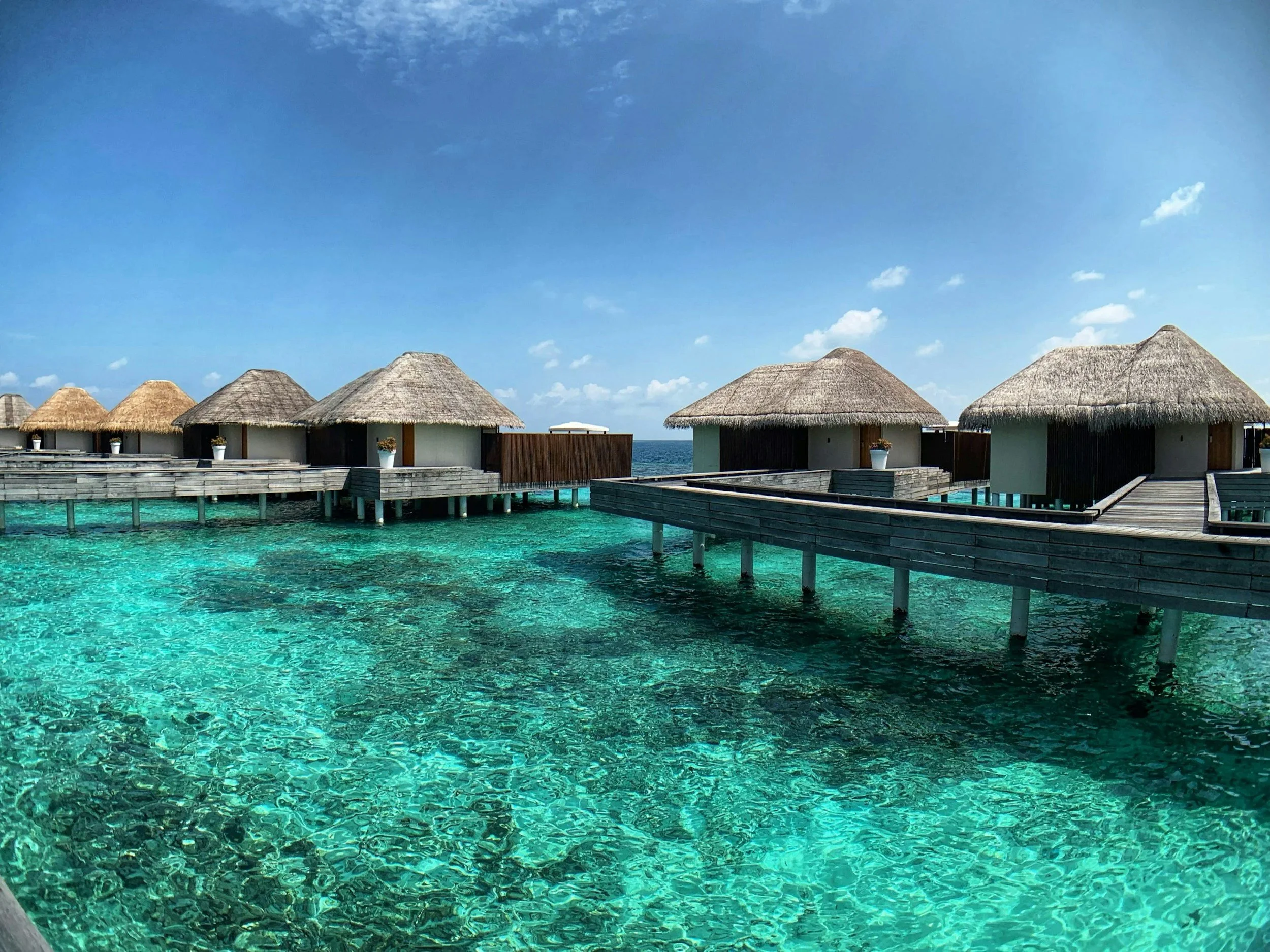 Overwater bungalows with thatched roofs and wooden walkways over clear turquoise ocean water on a sunny day.
