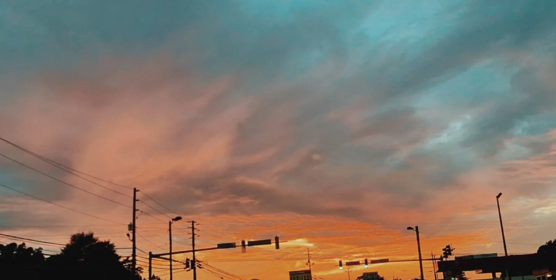 A vibrant sunset sky with colorful clouds, traffic lights, power lines, and streetlights over an urban street scene.