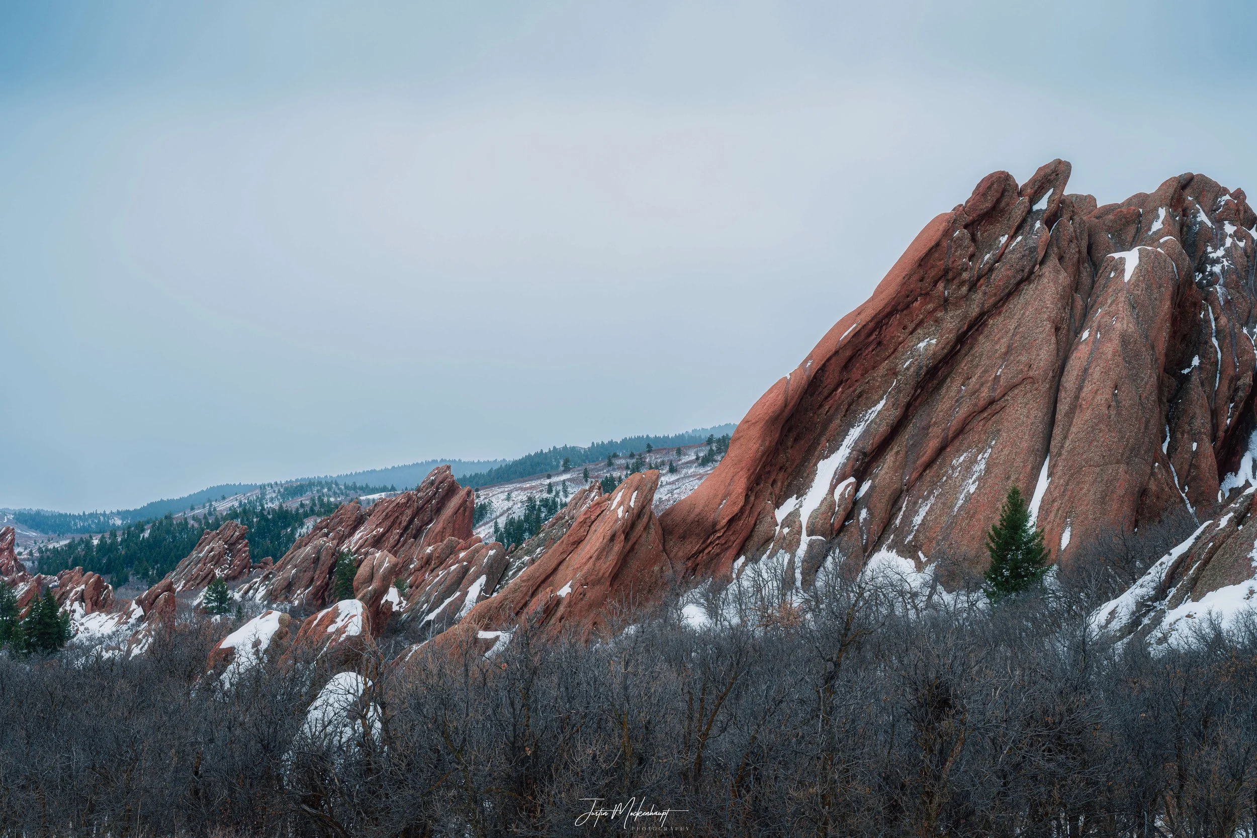 Snow-dusted red rock formations in a mountain landscape with dark leafless trees in the foreground and a forested hill in the background