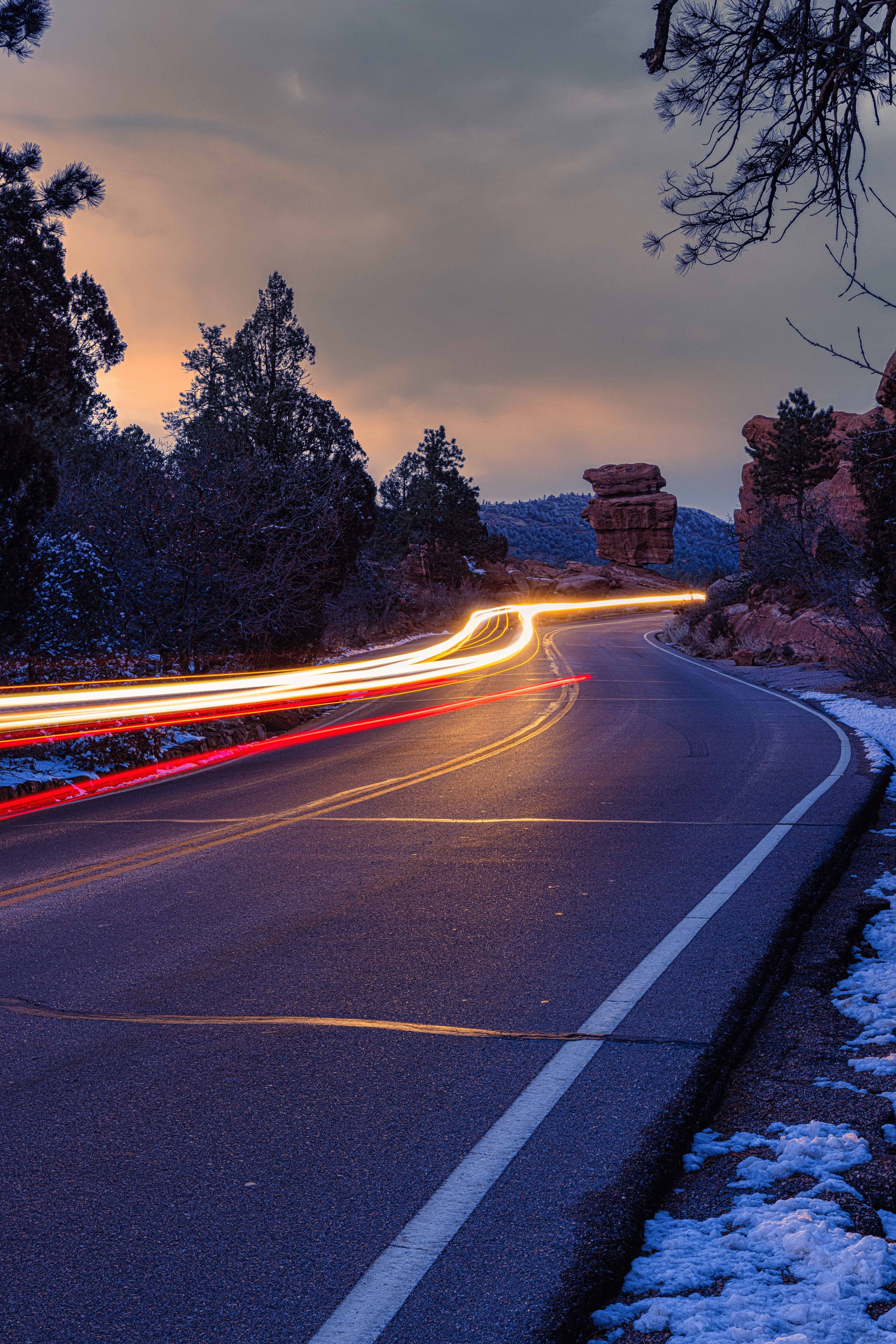 Winding mountain road at dusk with snow on the sides, trees on both sides, and light trails from passing vehicles.