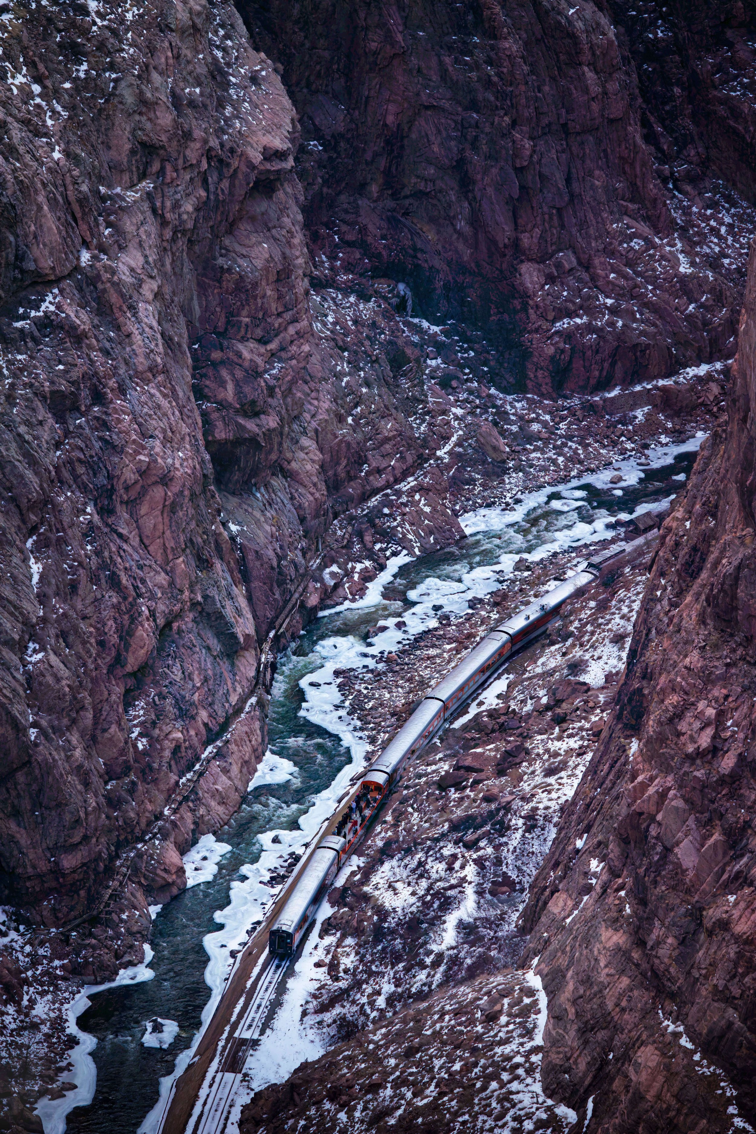 A mountain train traveling through a narrow canyon with rocky cliffs and patches of snow.