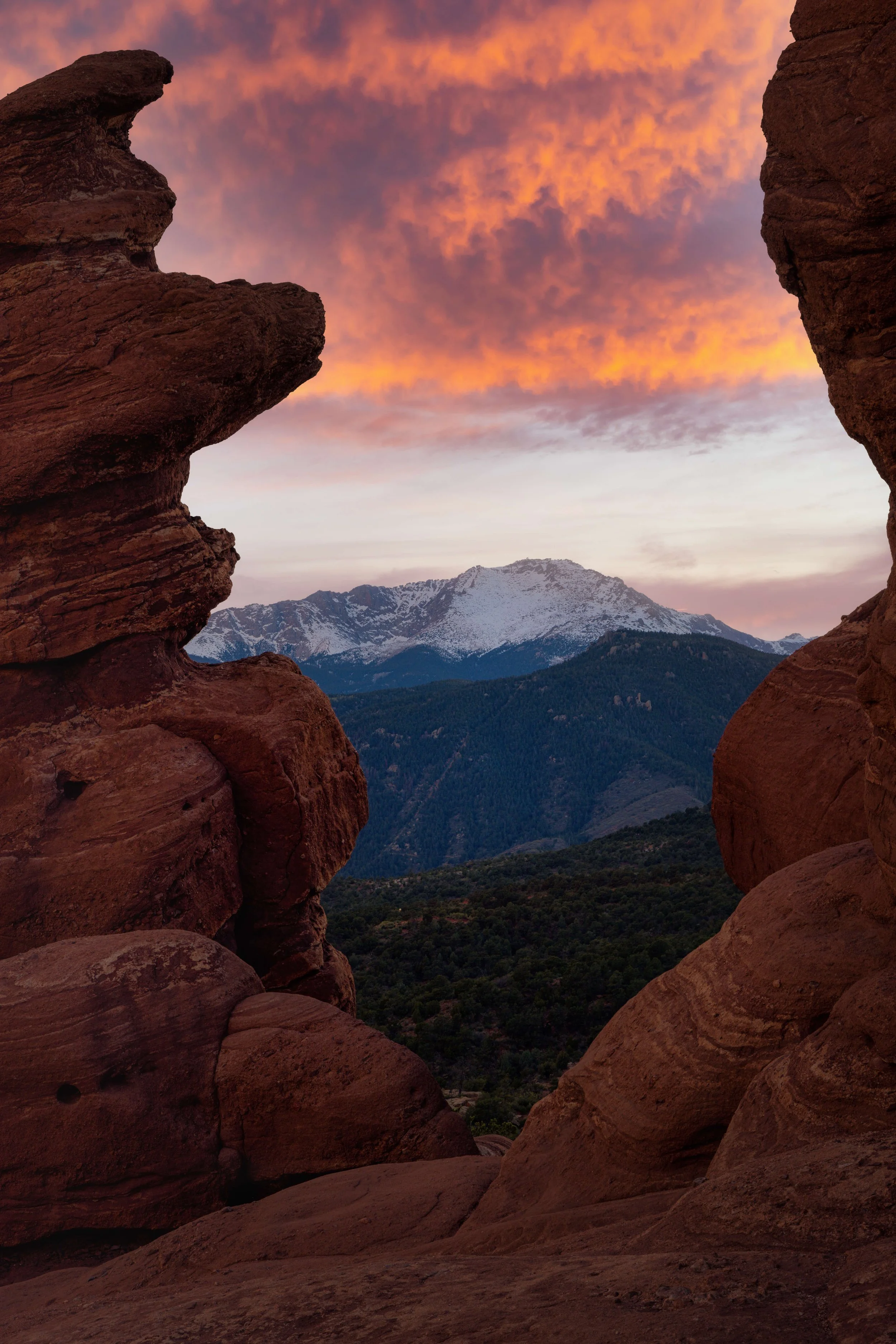 A view through a natural rock formation at sunset, showing snow-capped mountains and a colorful sky with orange clouds.