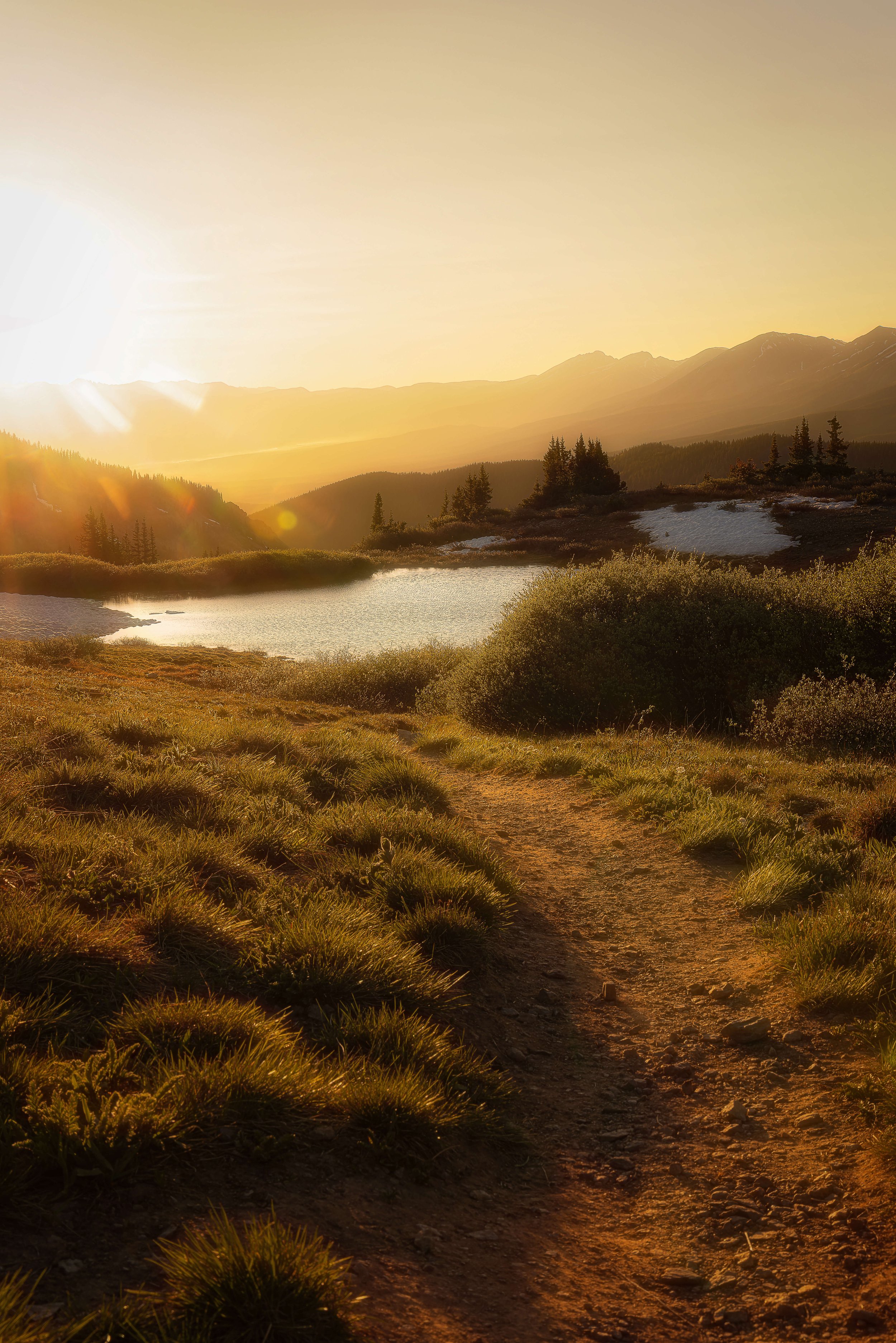 A scenic landscape of a mountain lake with a dirt trail, green bushes, and trees, highlighted by a setting or rising sun, with mountains in the background.