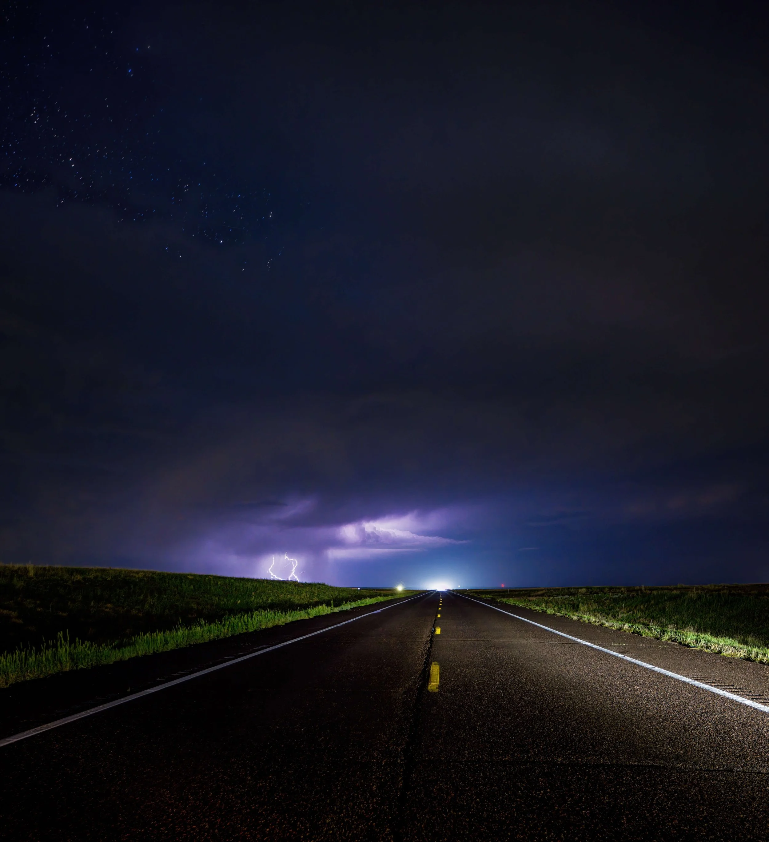 A long straight road with yellow dividing lines stretches into the distance under a dark, stormy sky with lightning, clouds, stars, and some distant light