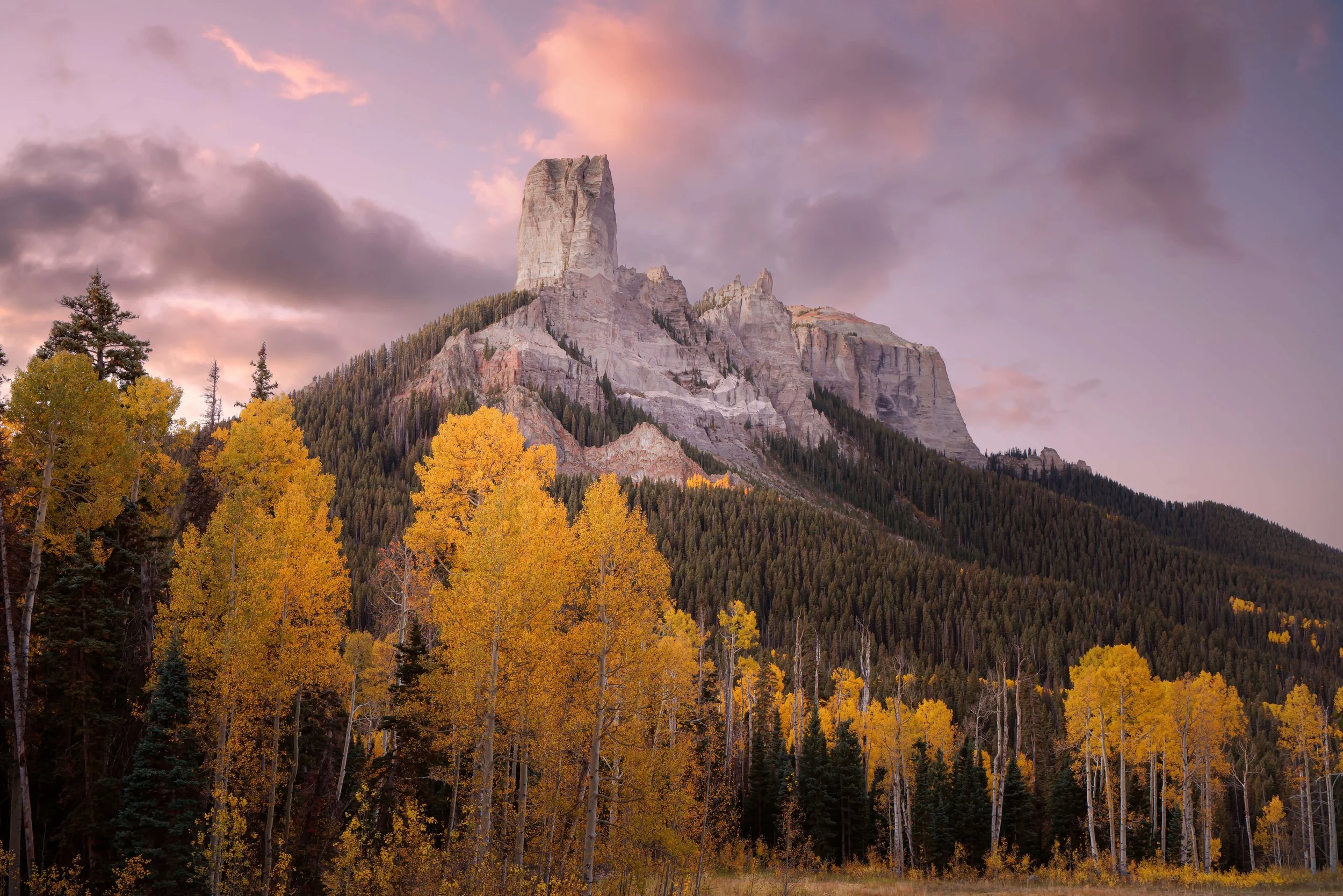 A mountain with a tall, flat top surrounded by forested slopes and colorful autumn trees in the foreground at sunset.