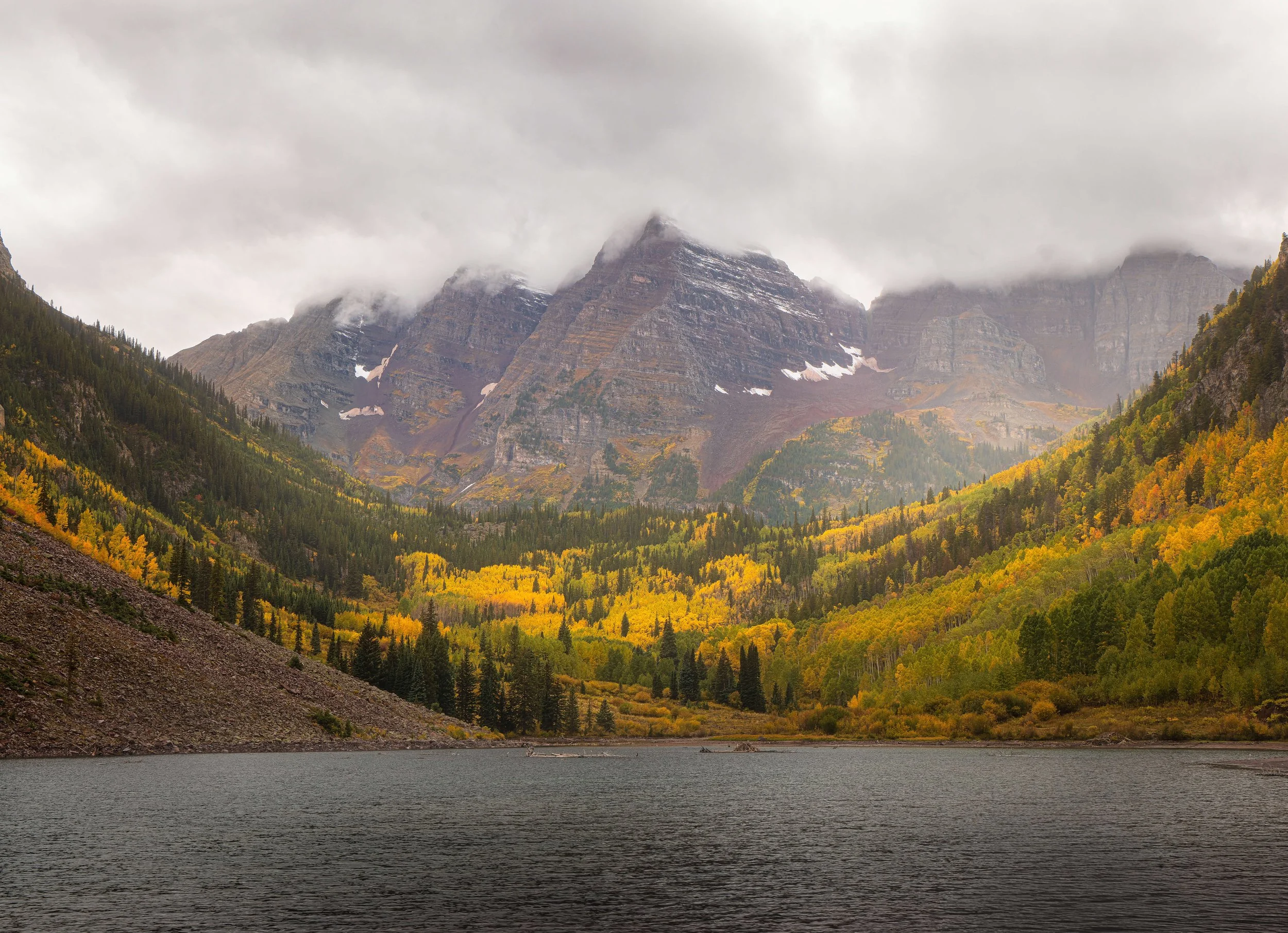 A mountain landscape with a cloudy sky, dense green and yellow trees in the valley, and a body of water in the foreground.