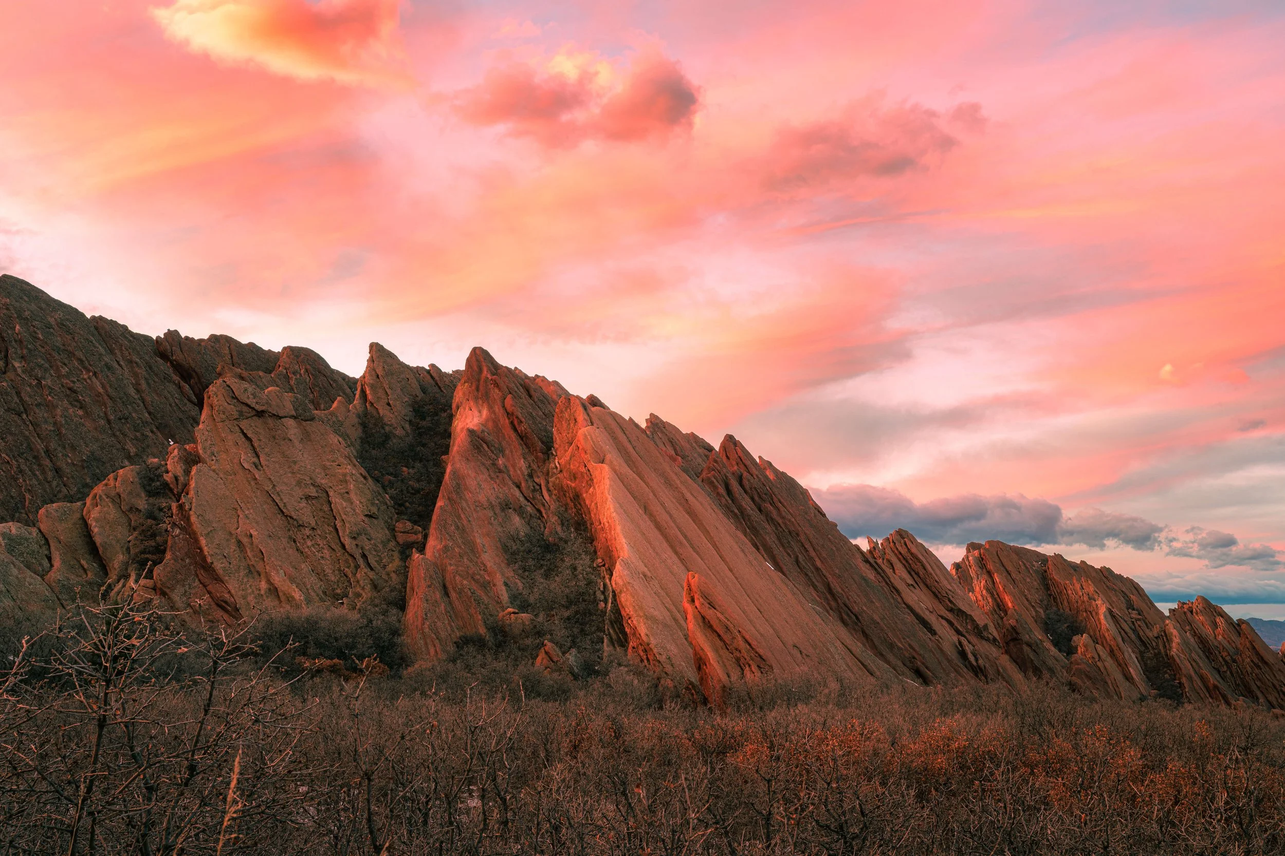 Sunset sky over rugged mountain peaks with pink and orange clouds and sparse vegetation in the foreground.