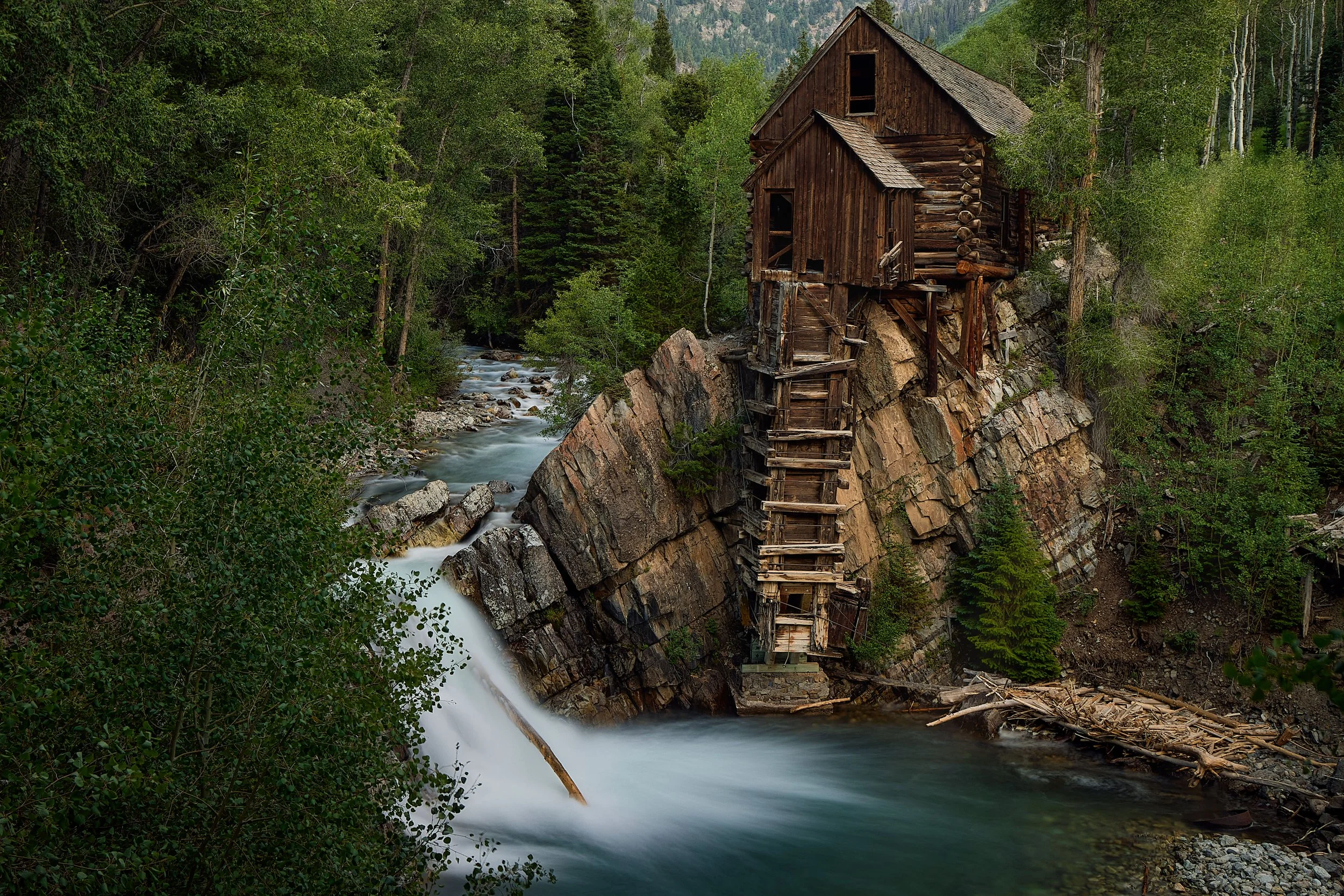 Old wooden house built on a rocky cliff overlooking a flowing river in a dense forest.