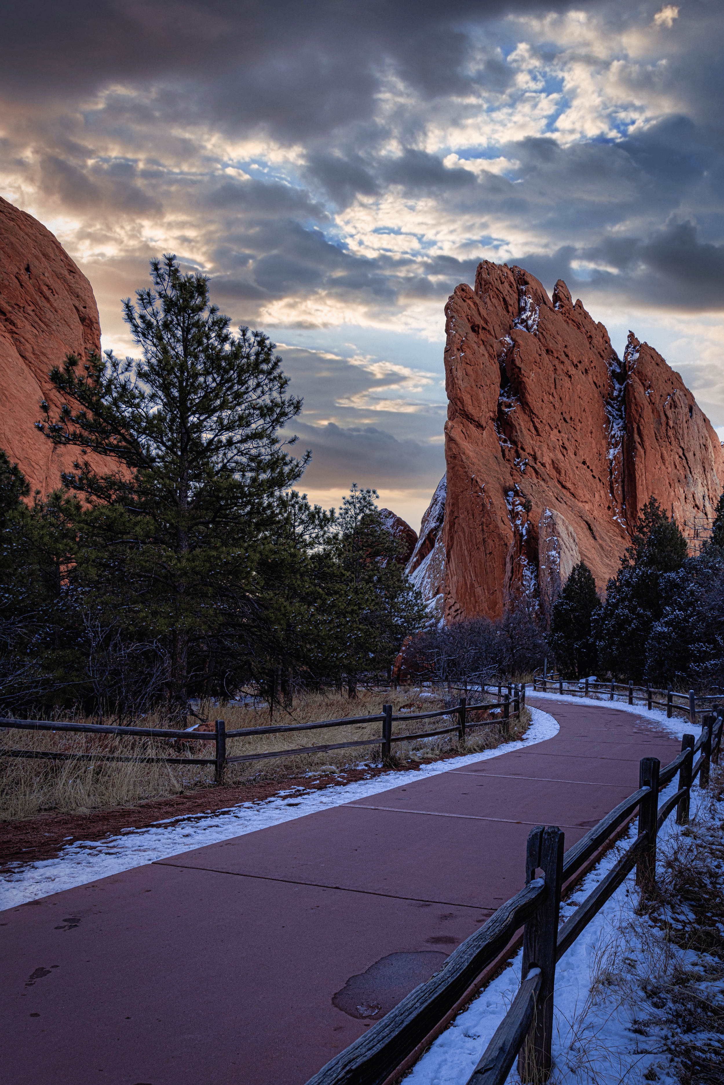 A winding pathway through a snowy landscape with tall red rock formations and pine trees under a cloudy sky.