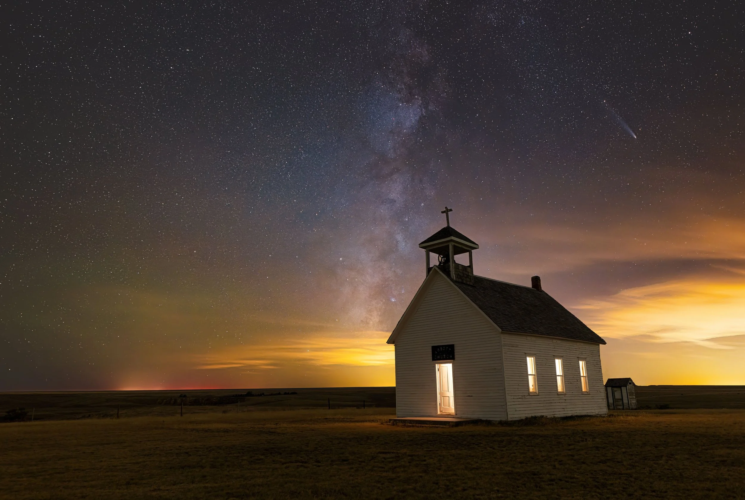 A small white church building with illuminated windows under a night sky filled with stars and the Milky Way, with the horizon showing a glow of city lights or sunset.