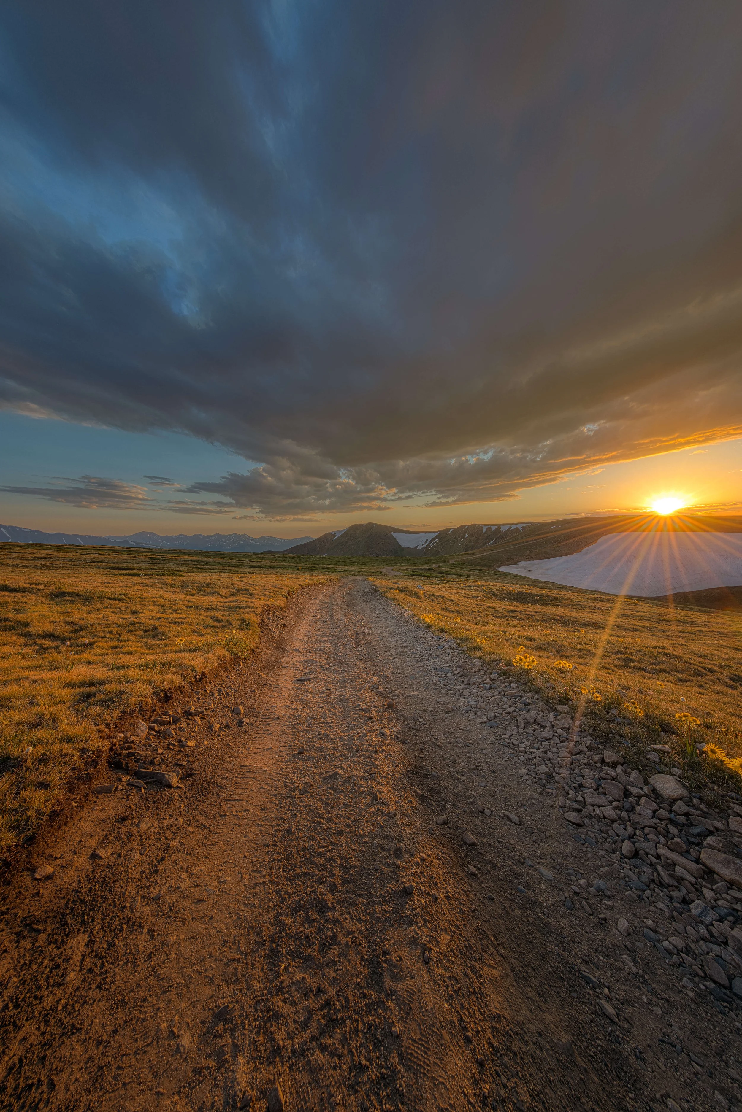 A dirt trail running through a grassy plain during sunset with mountain ranges in the background and dark clouds in the sky.