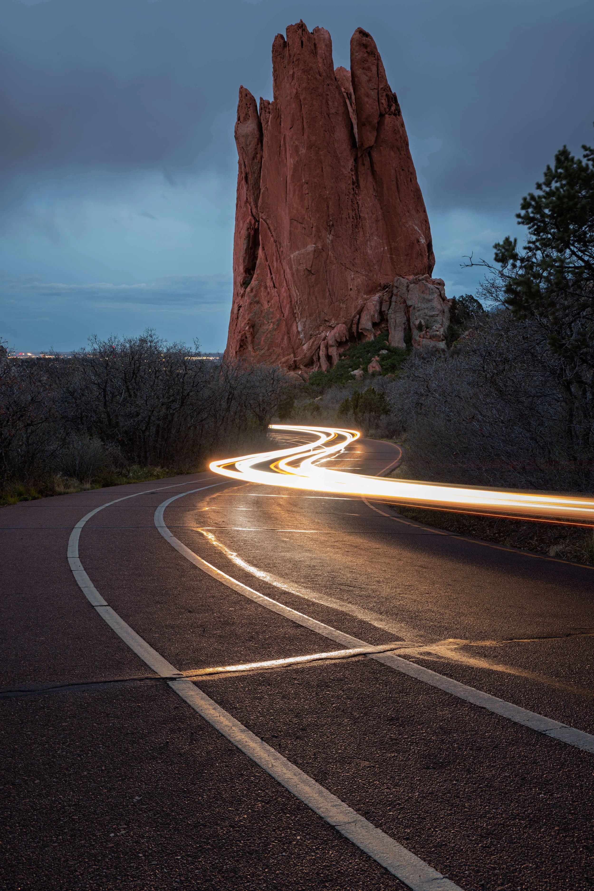 A winding road at dusk with car light trails leading towards a large red rock formation and dark cloudy sky.