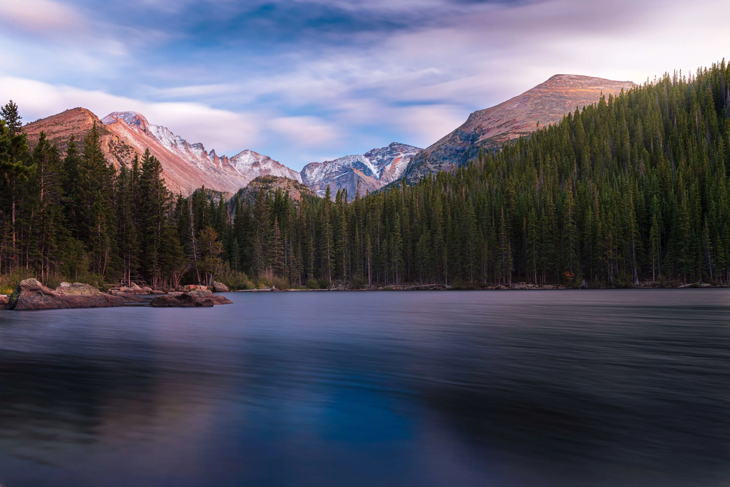 A scenic view of a mountain lake with a dense forest of pine trees, and rugged mountains with some snow on top in the background under a partly cloudy sky.