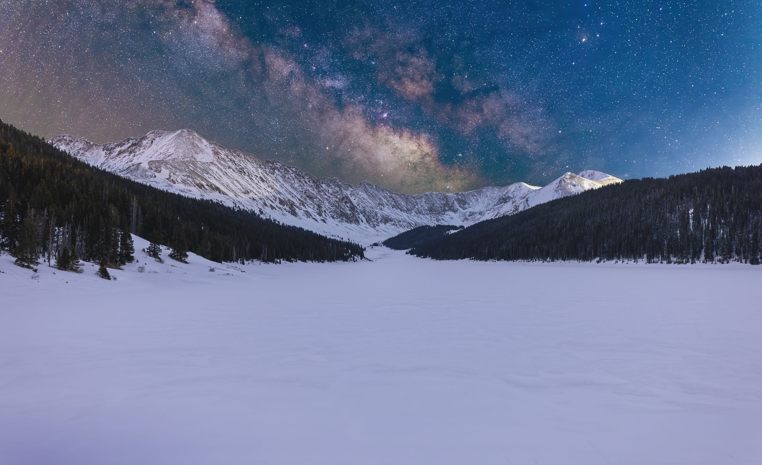 Snow-covered valley surrounded by forested mountains under a starry night sky with the Milky Way galaxy visible.