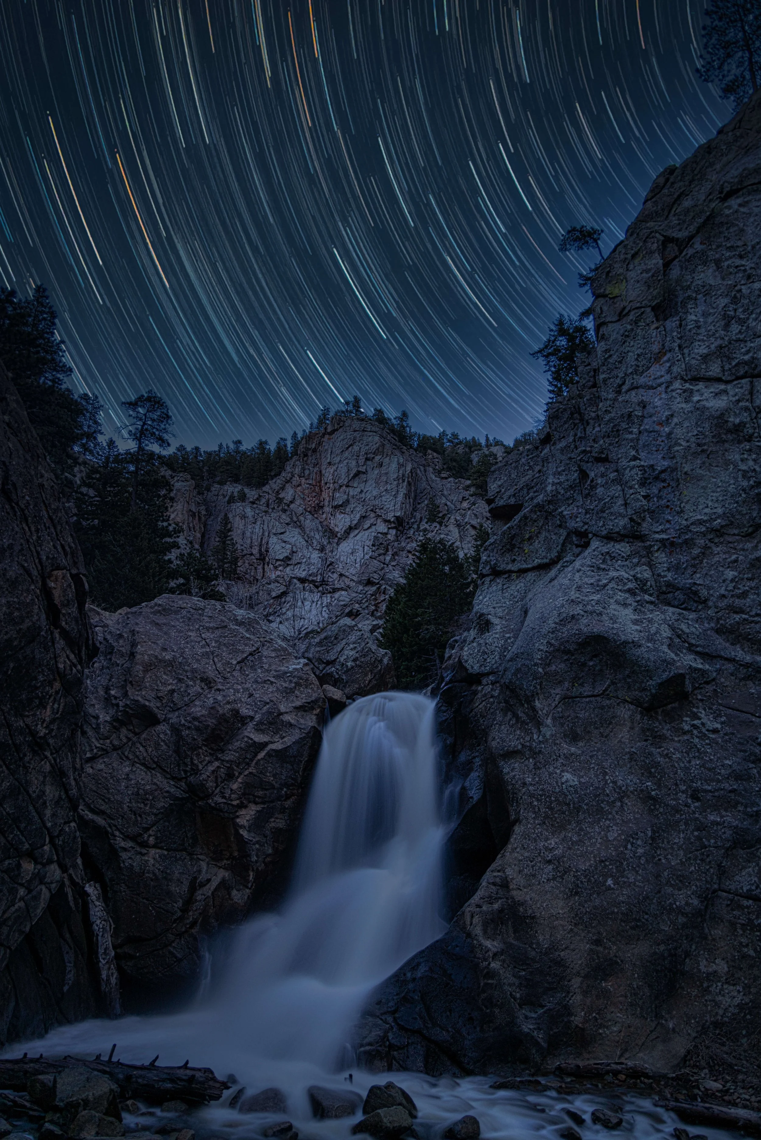 Nighttime landscape of a rocky canyon with a small waterfall and tall trees, under a star-filled sky with star trails.