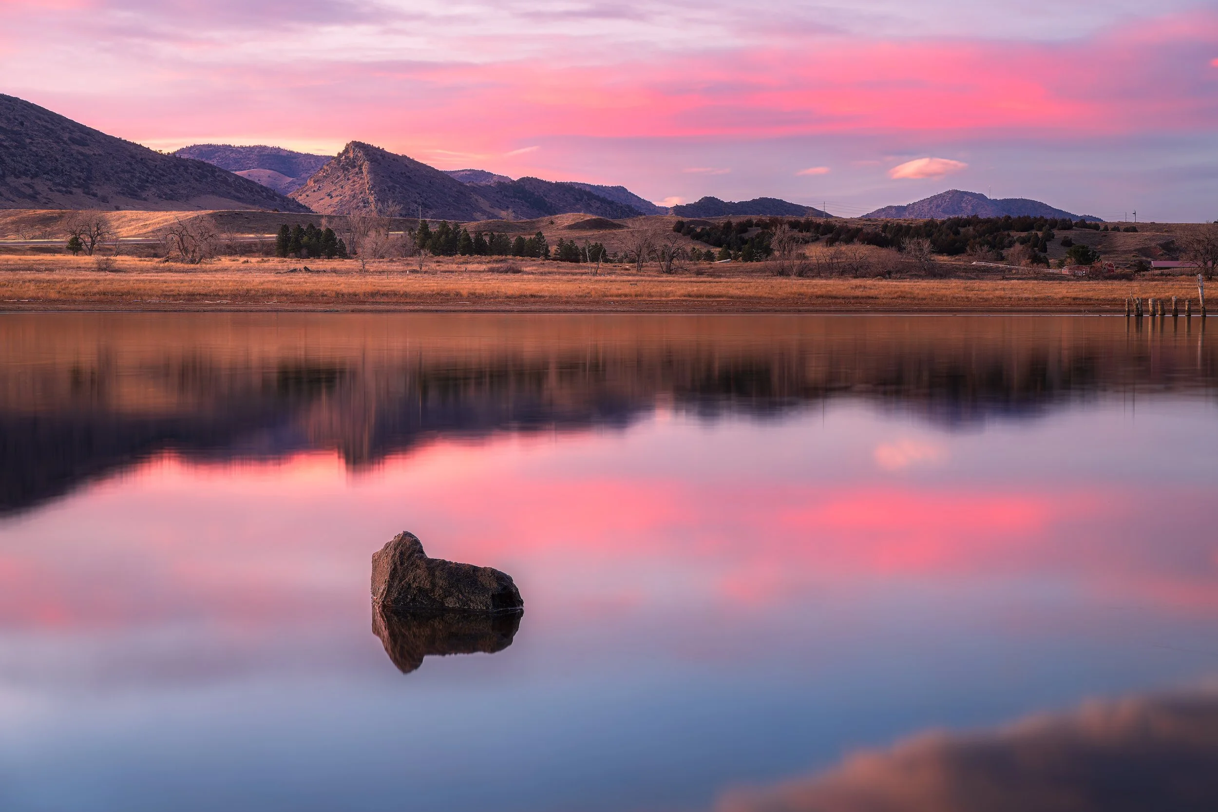 Scenic landscape with a calm lake reflecting a colorful pink and purple sunset sky, surrounded by mountains and sparse trees.