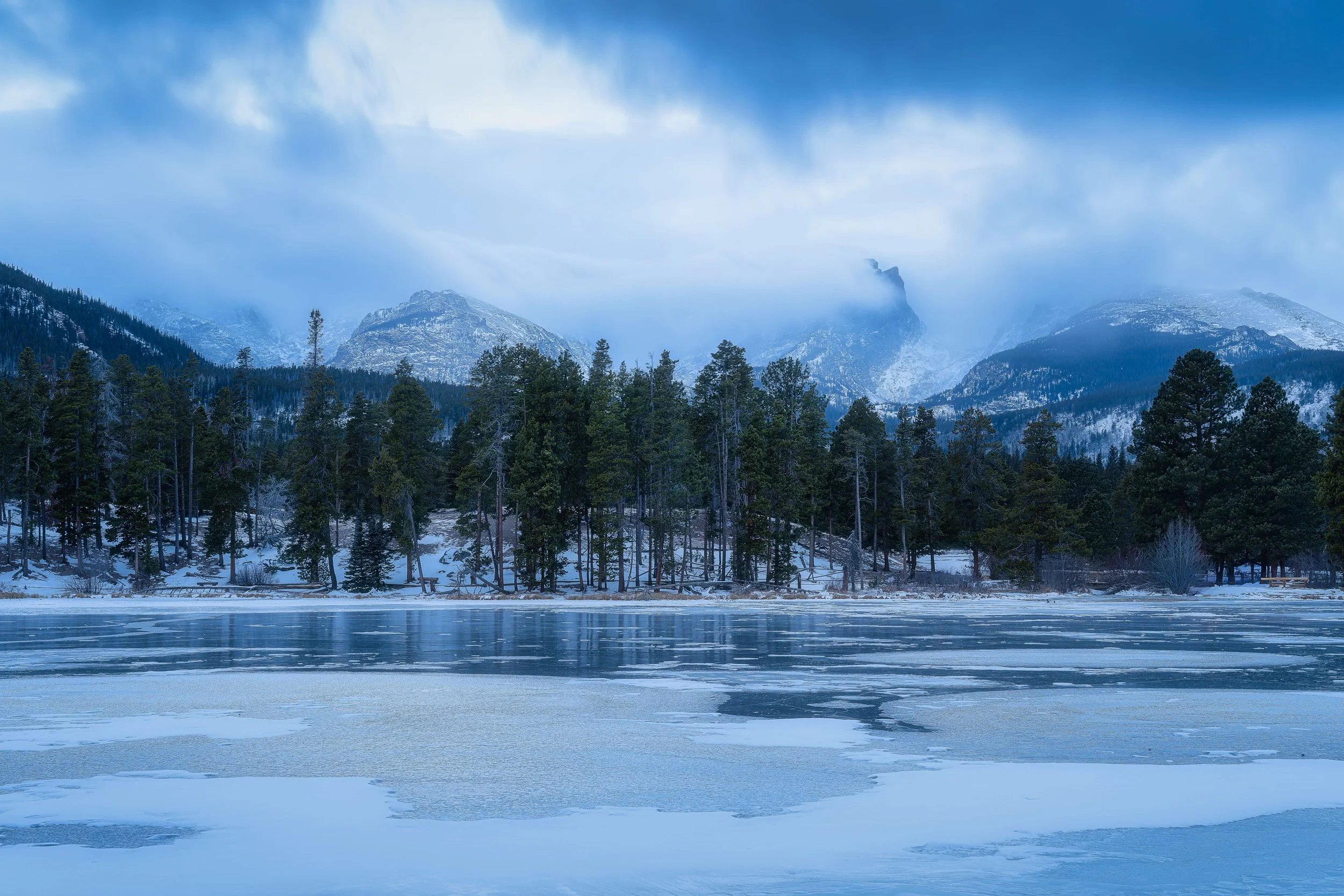 A winter landscape featuring a partially frozen lake with snow and ice, dense evergreen trees along the shoreline, and tall mountains in the background, surrounded by clouds and mist.