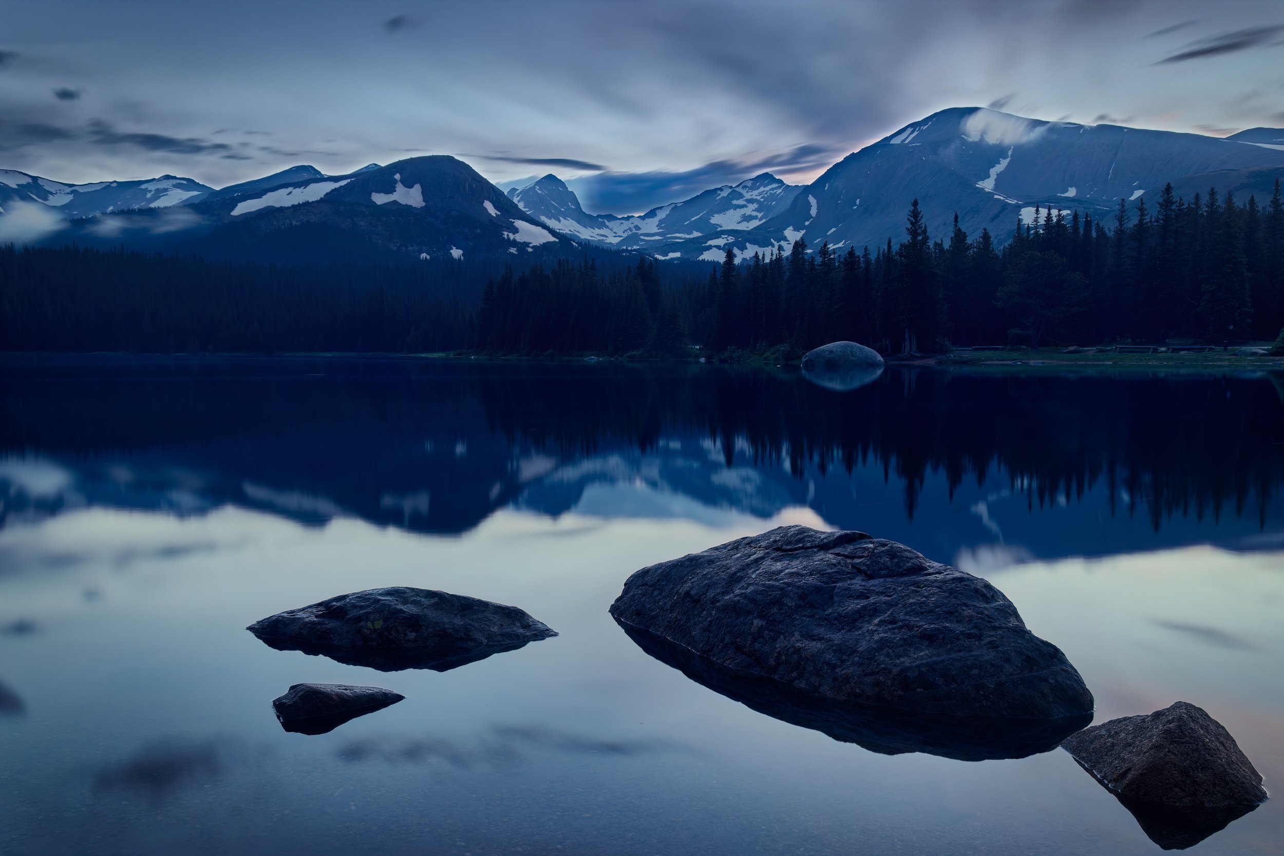 Mountain lake scene with large rocks in the foreground, calm water reflecting surrounding pine trees, mountains with patches of snow, and a cloudy sky.