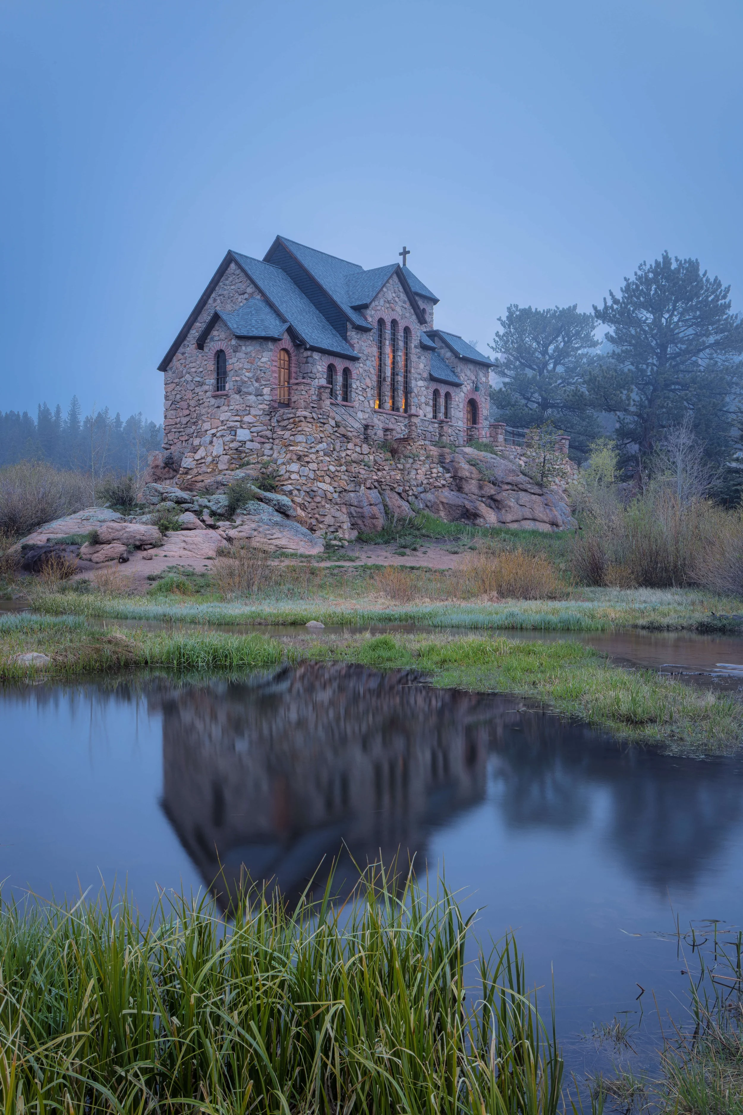 A stone church with a cross on top, situated on a rocky hill surrounded by trees, with a pond reflecting the church in the foreground.