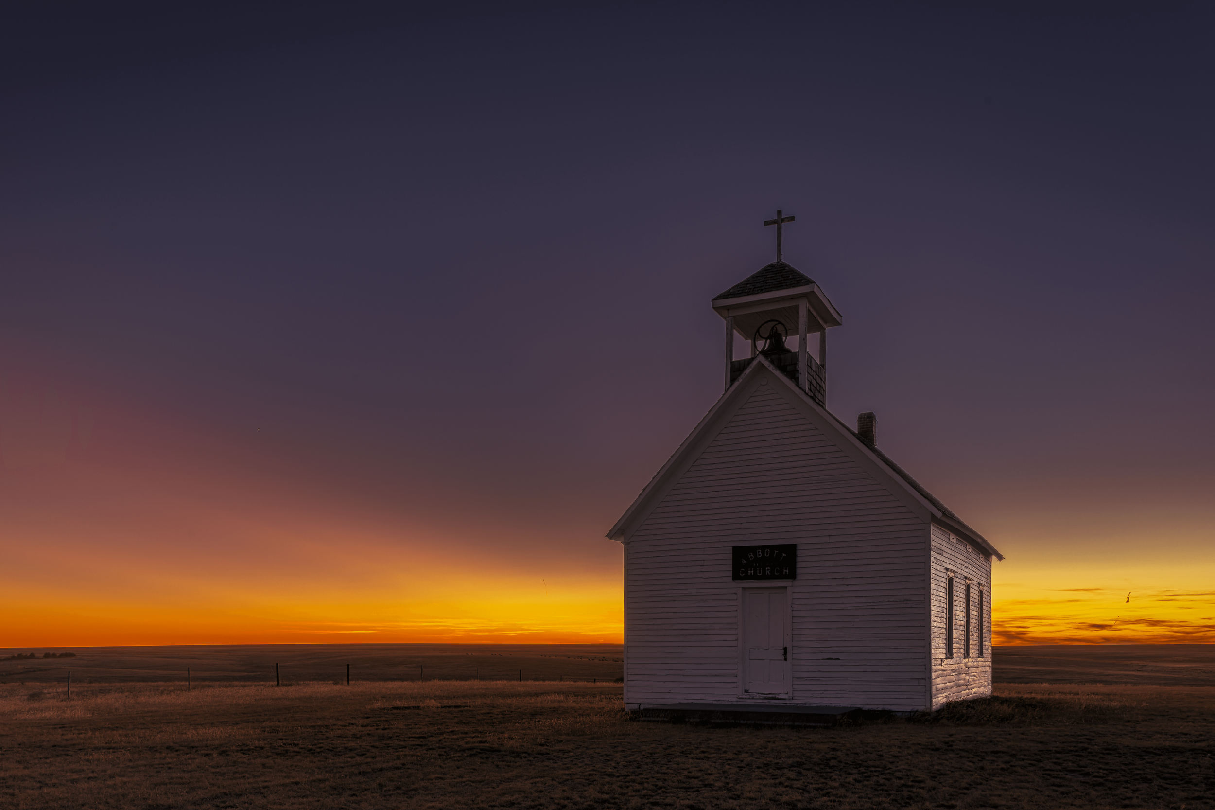 Small white church with a steeple and cross on top, located in a vast open field at sunset with a colorful sky