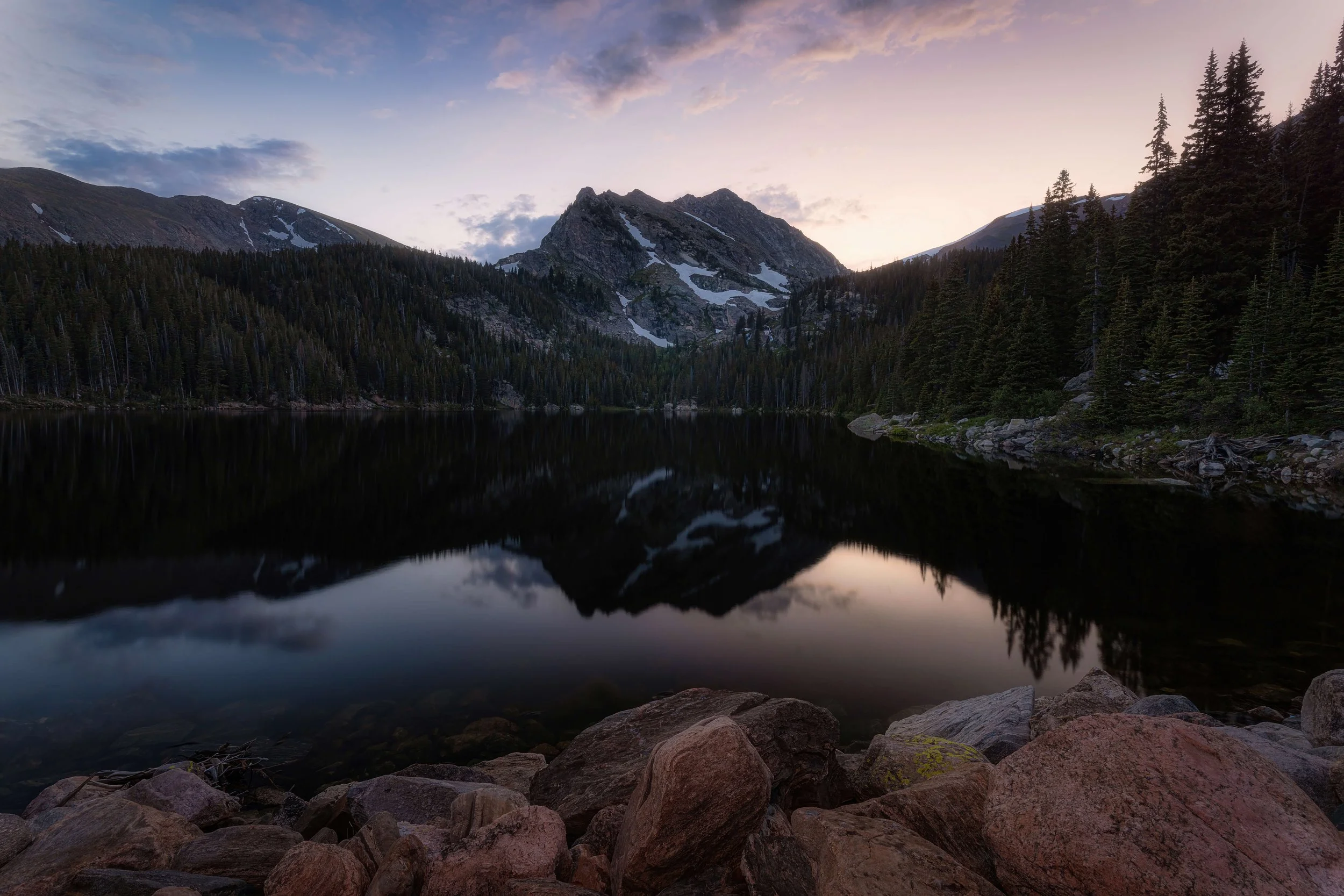 A serene mountain lake at dusk, surrounded by dense evergreen trees and rocky shoreline, with snow-capped peaks reflected in the calm water, under a pastel-colored sky.