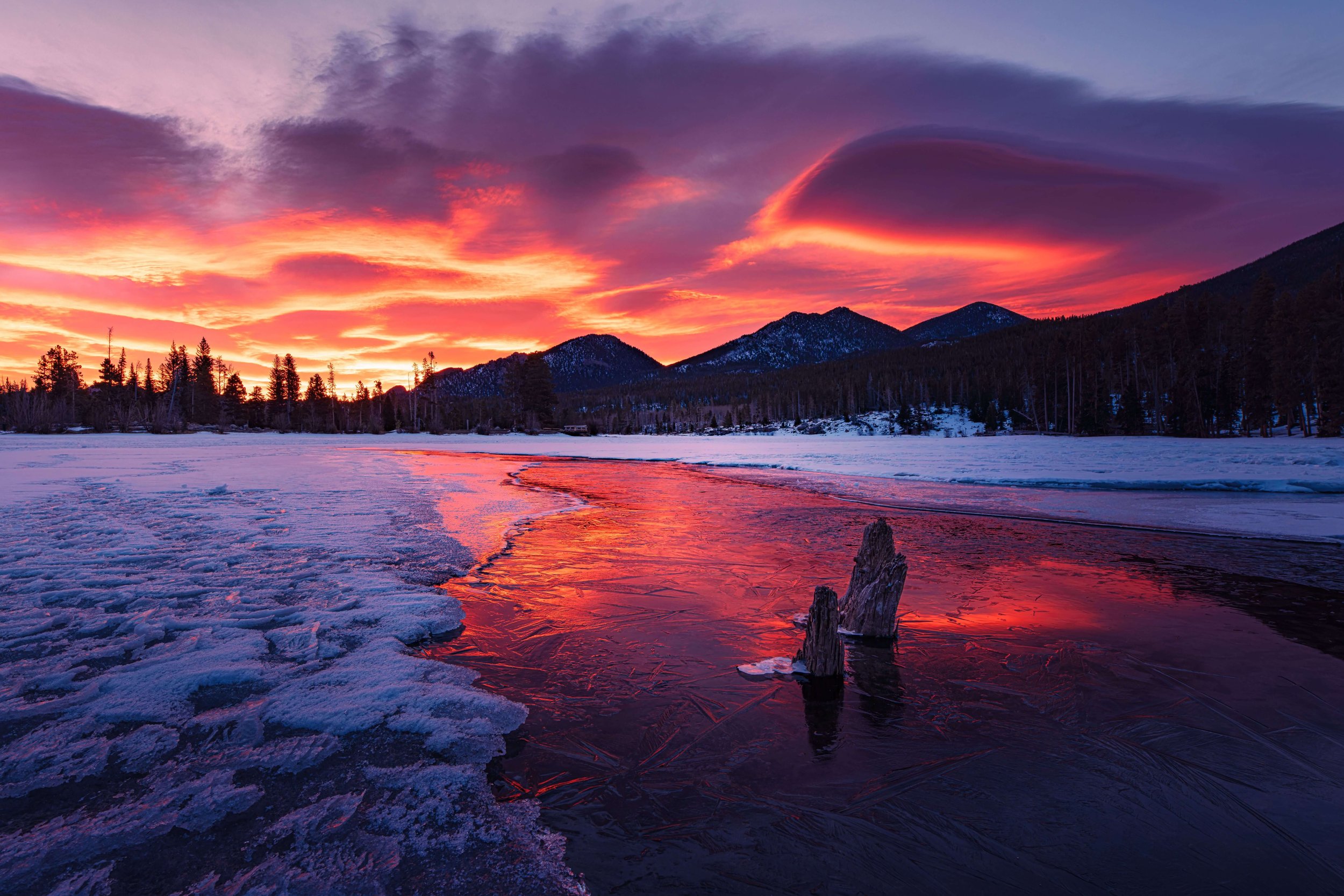 Snow-covered landscape with a partially frozen river reflecting a vibrant sunset sky, mountains, and trees in the background.
