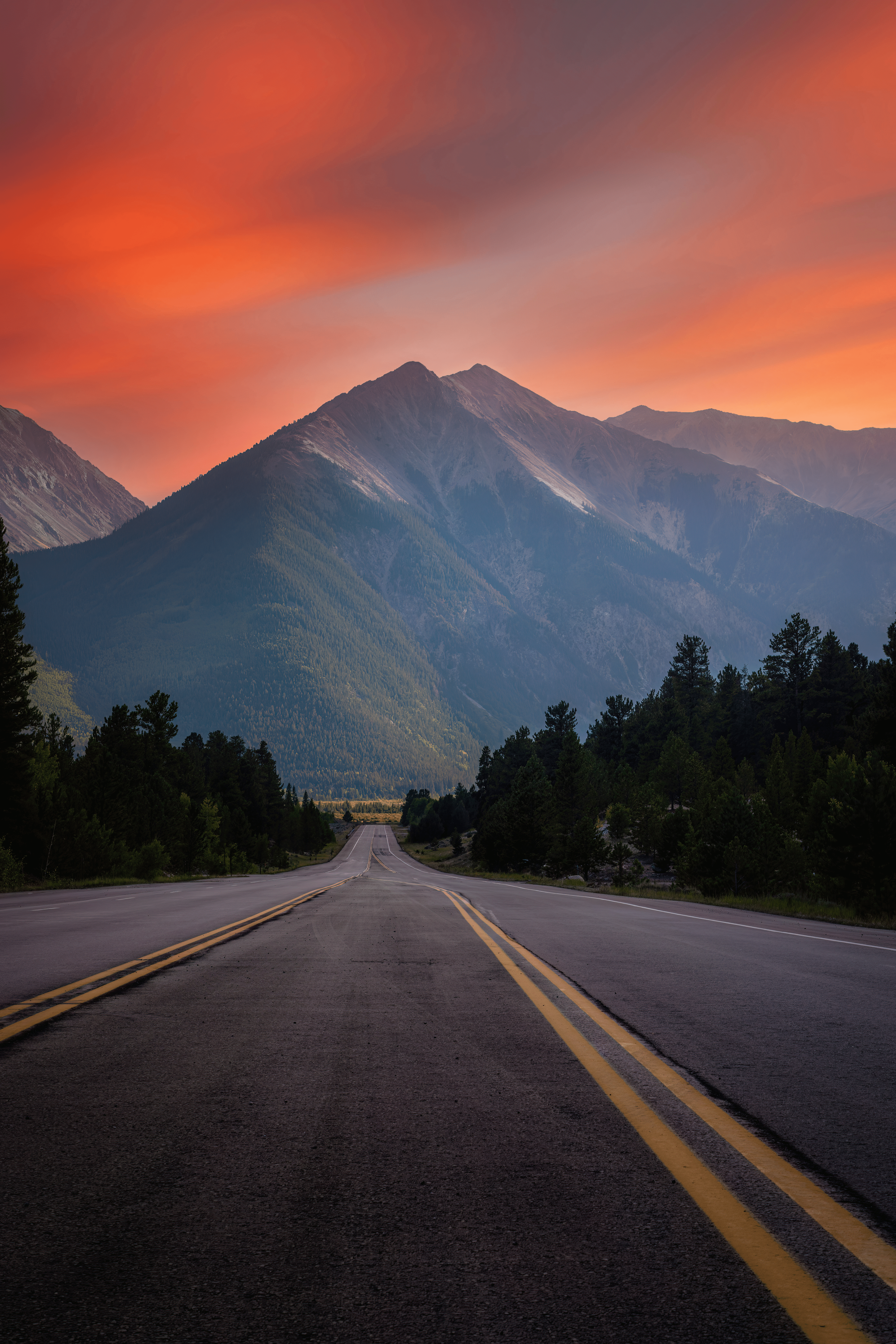 A two-lane road leading towards tall mountains with a sunset sky filled with orange and purple clouds, surrounded by dense forest.