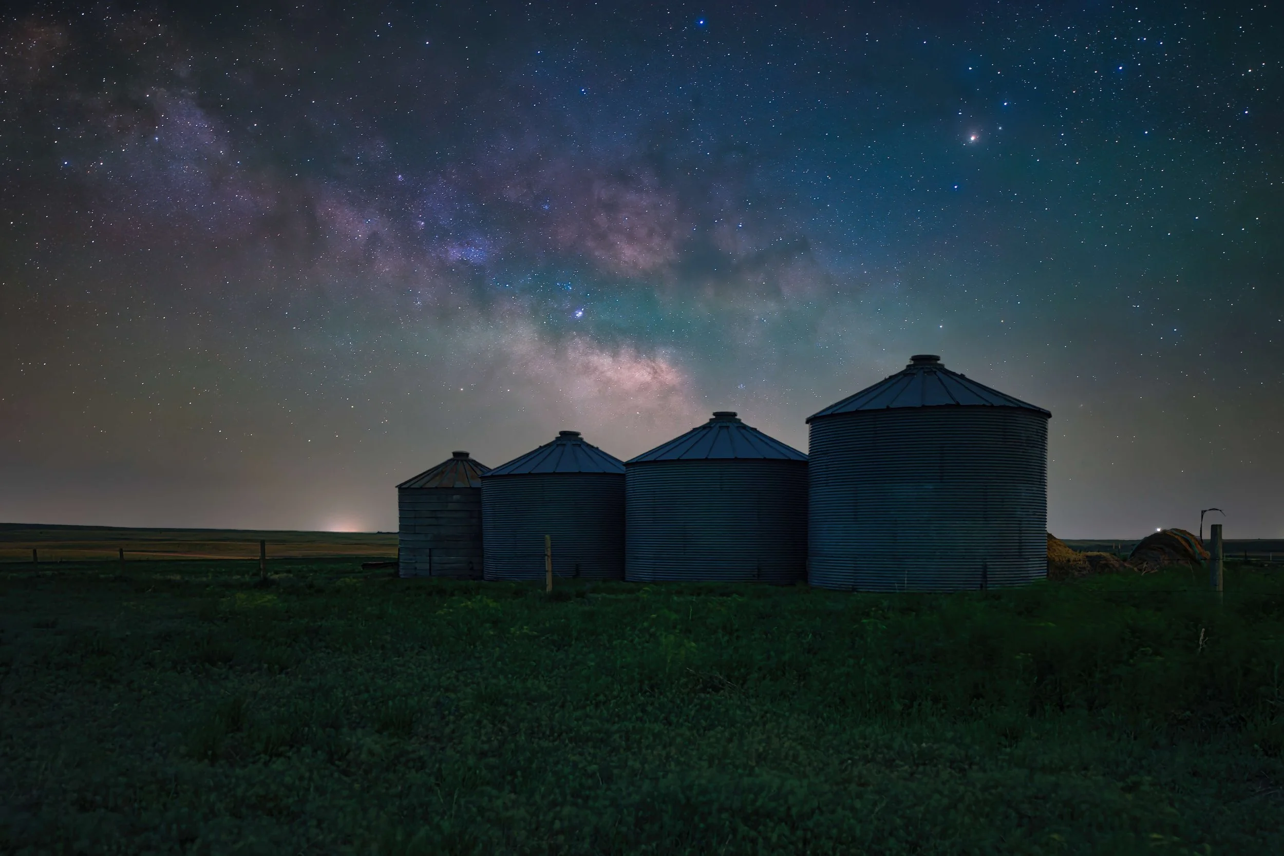 Night sky filled with stars and the Milky Way galaxy above four metal grain silos in a rural area.
